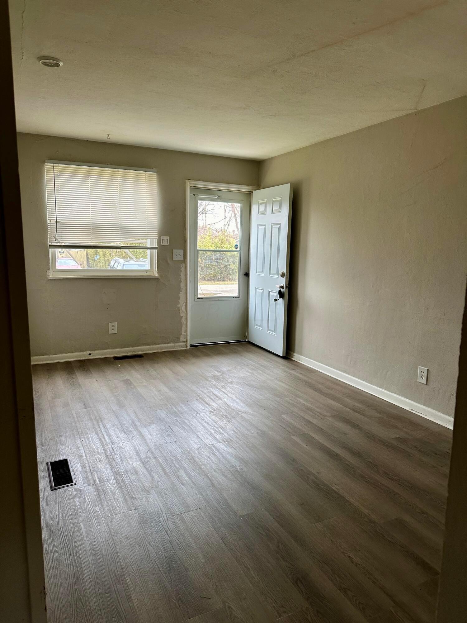 Empty room with gray wood-look flooring, a window with blinds, and an open front door.