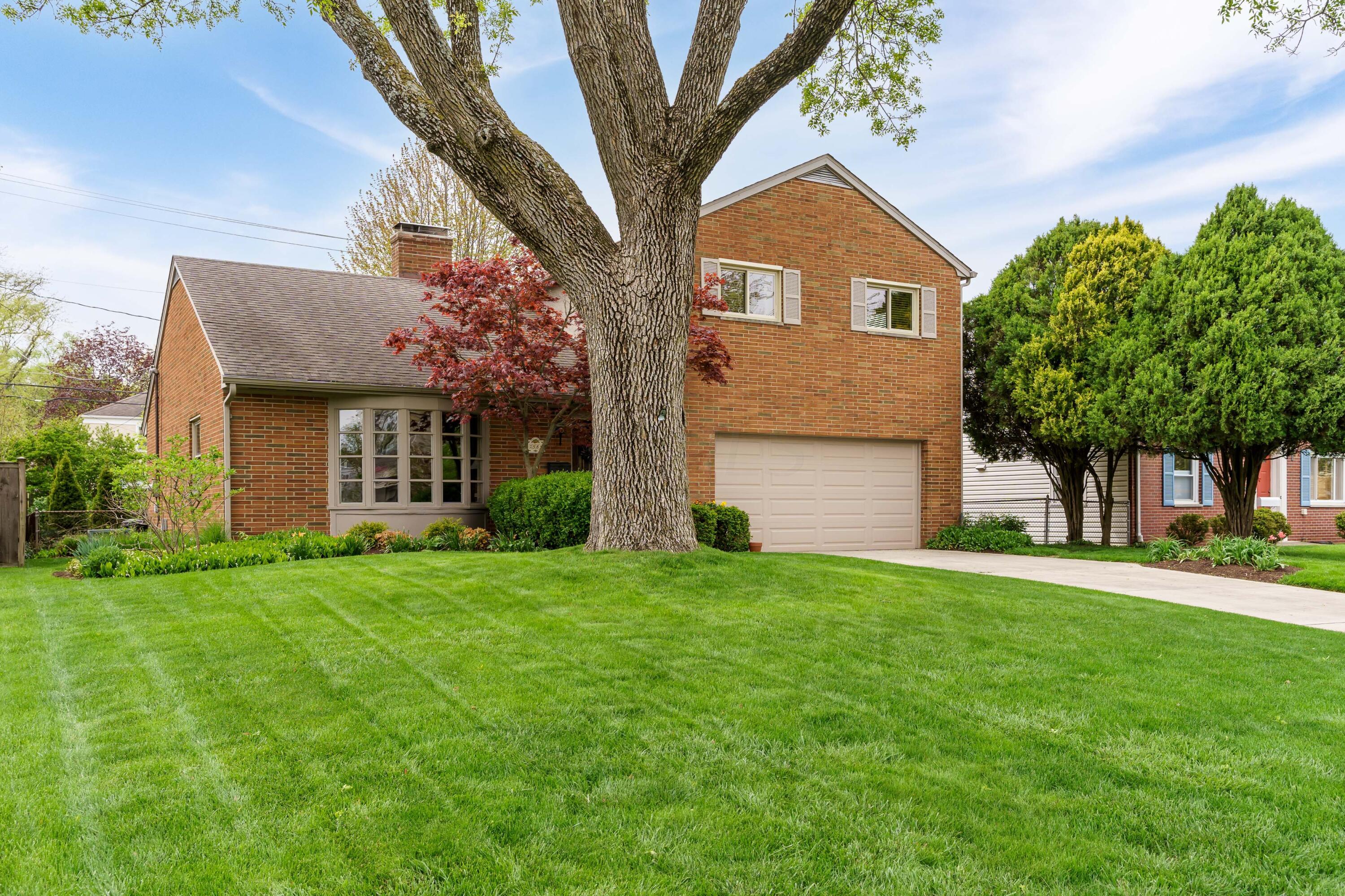 Two-story brick house with an attached garage, a large front-yard tree, and a broad green lawn.