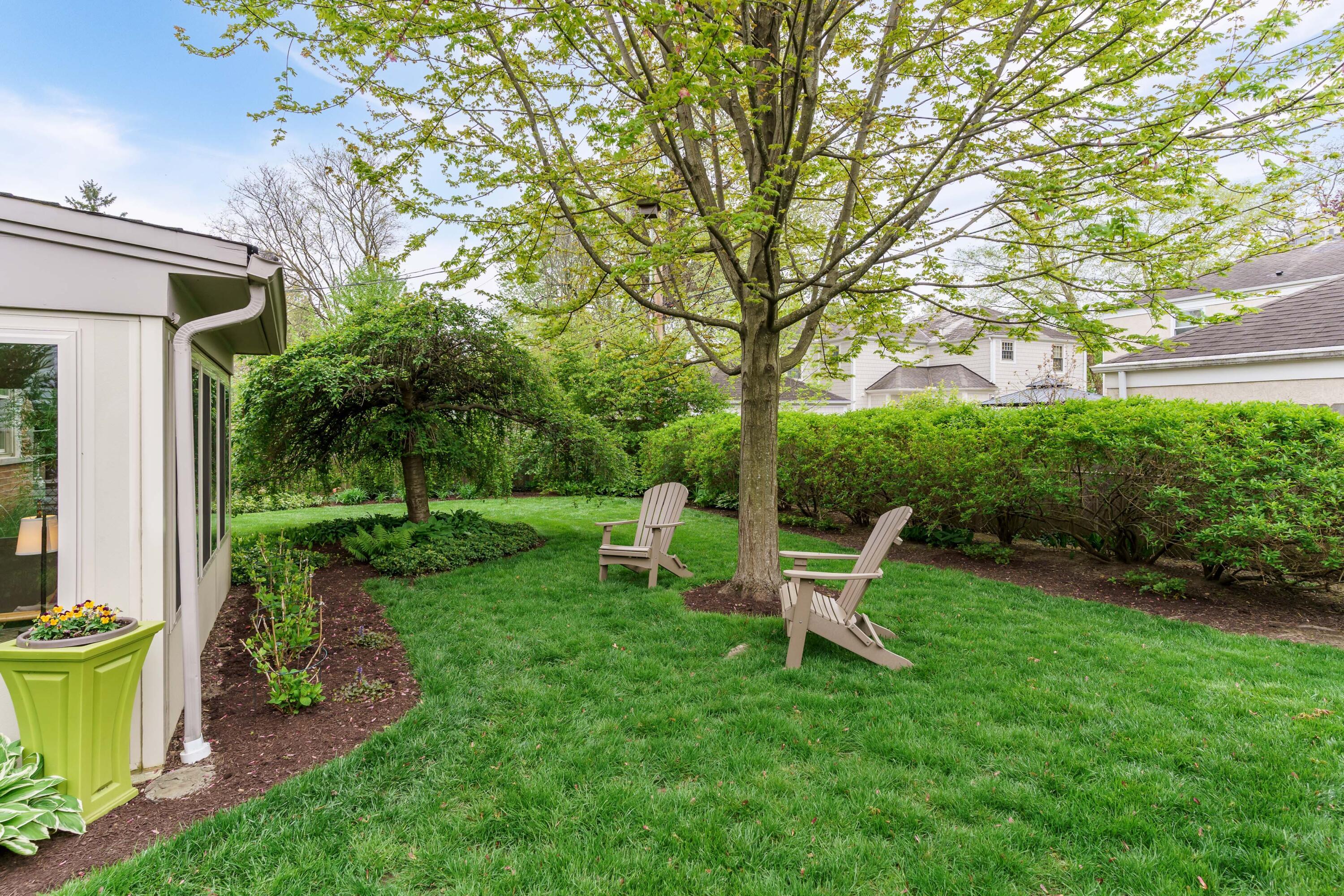 Landscaped backyard with a large shade tree, green lawn, and two Adirondack chairs.