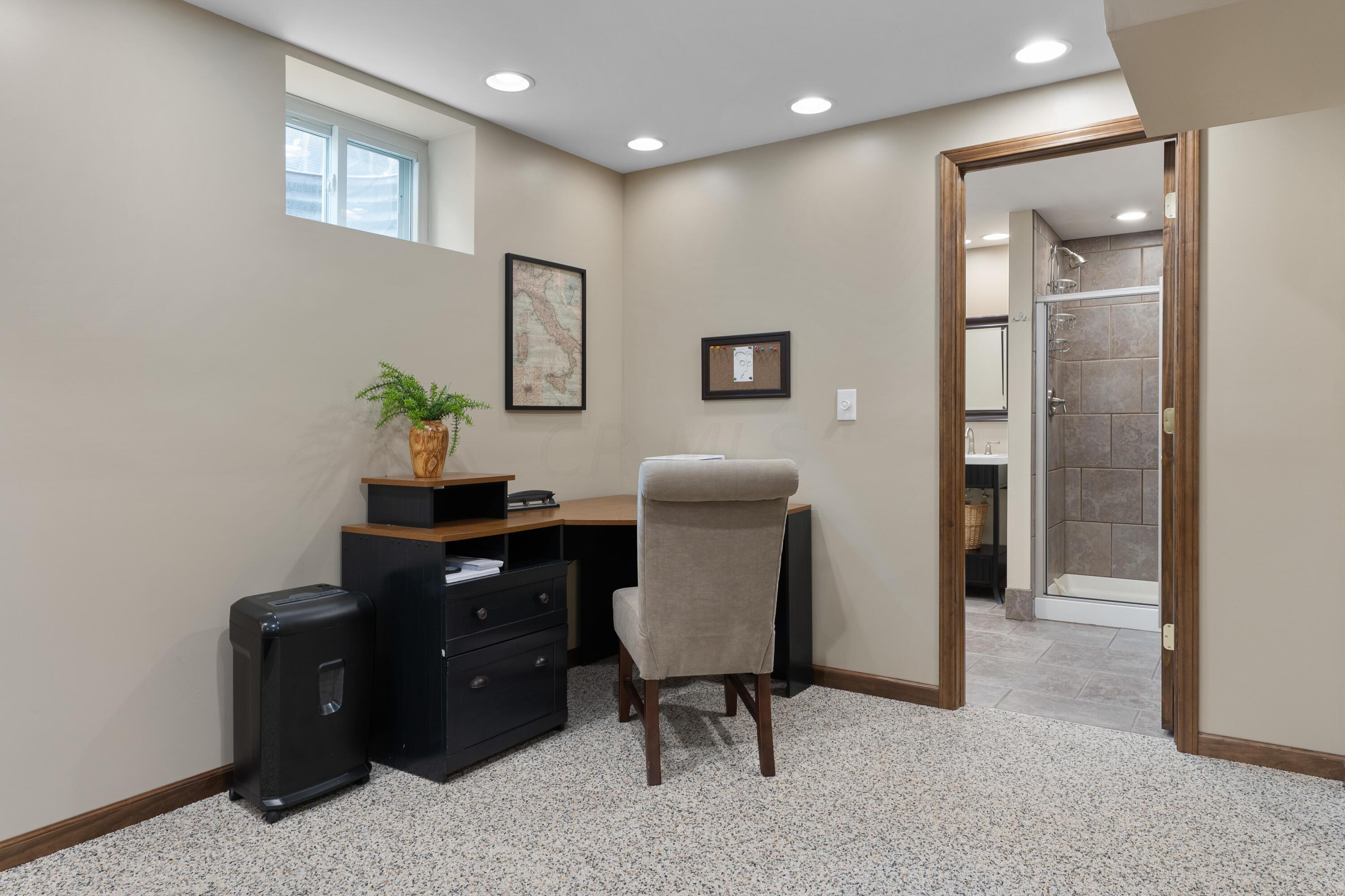Furnished home office with a dark desk and chair, opening to a tiled bathroom with a glass shower.
