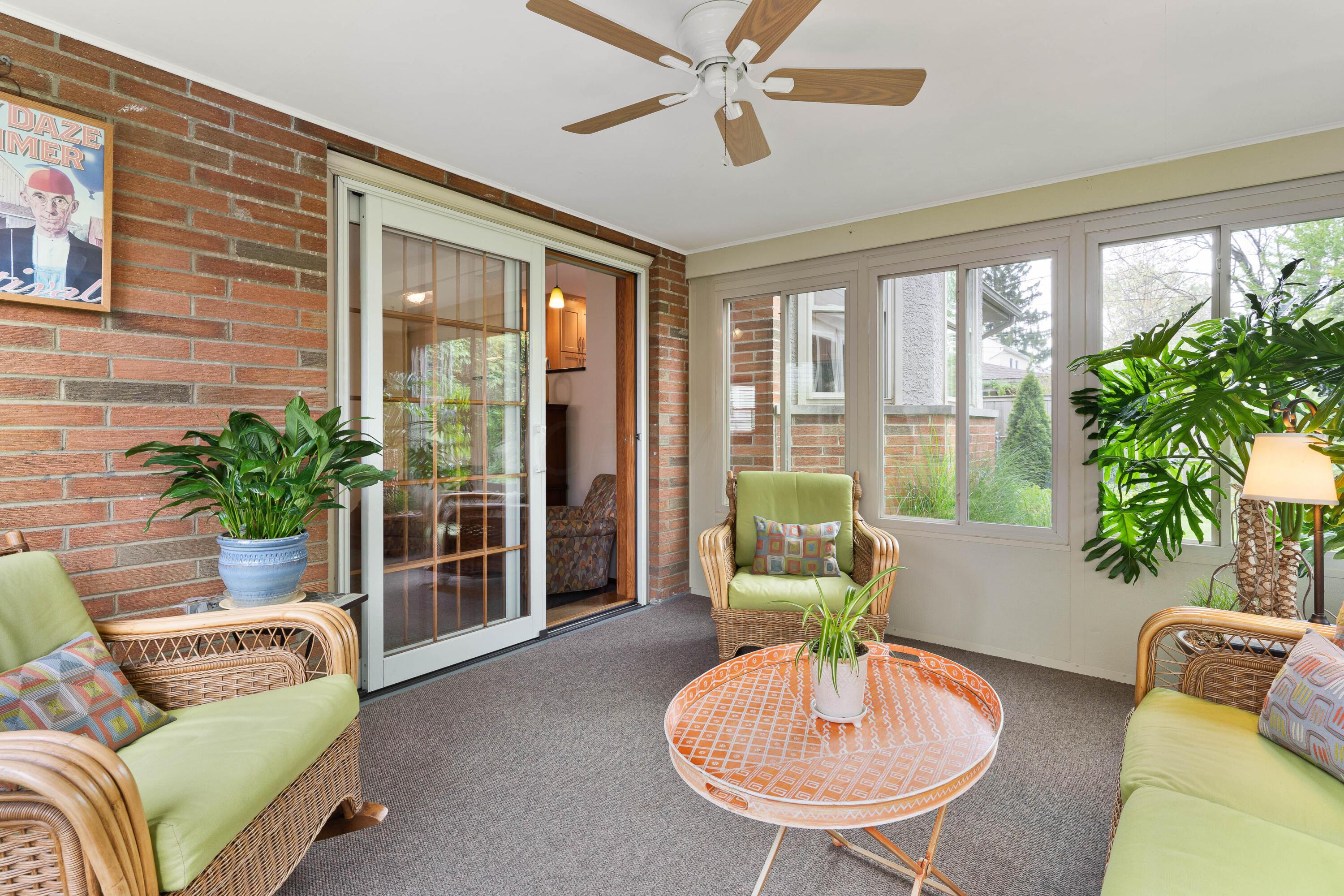 Sunroom with green-cushioned wicker chairs, an orange round table, brick wall, and large windows.