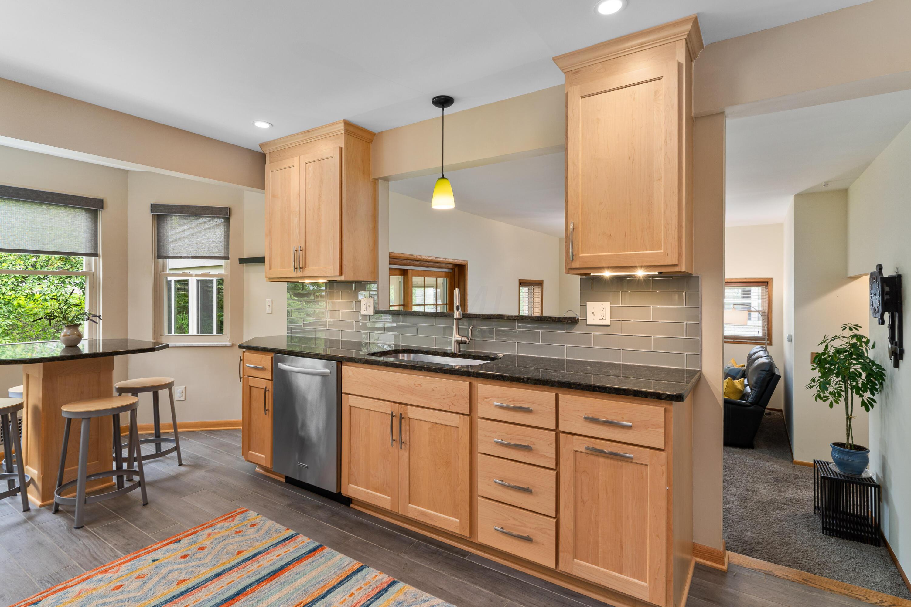Kitchen with light wood cabinets, dark counters, gray tile backsplash, and an adjoining sitting area.