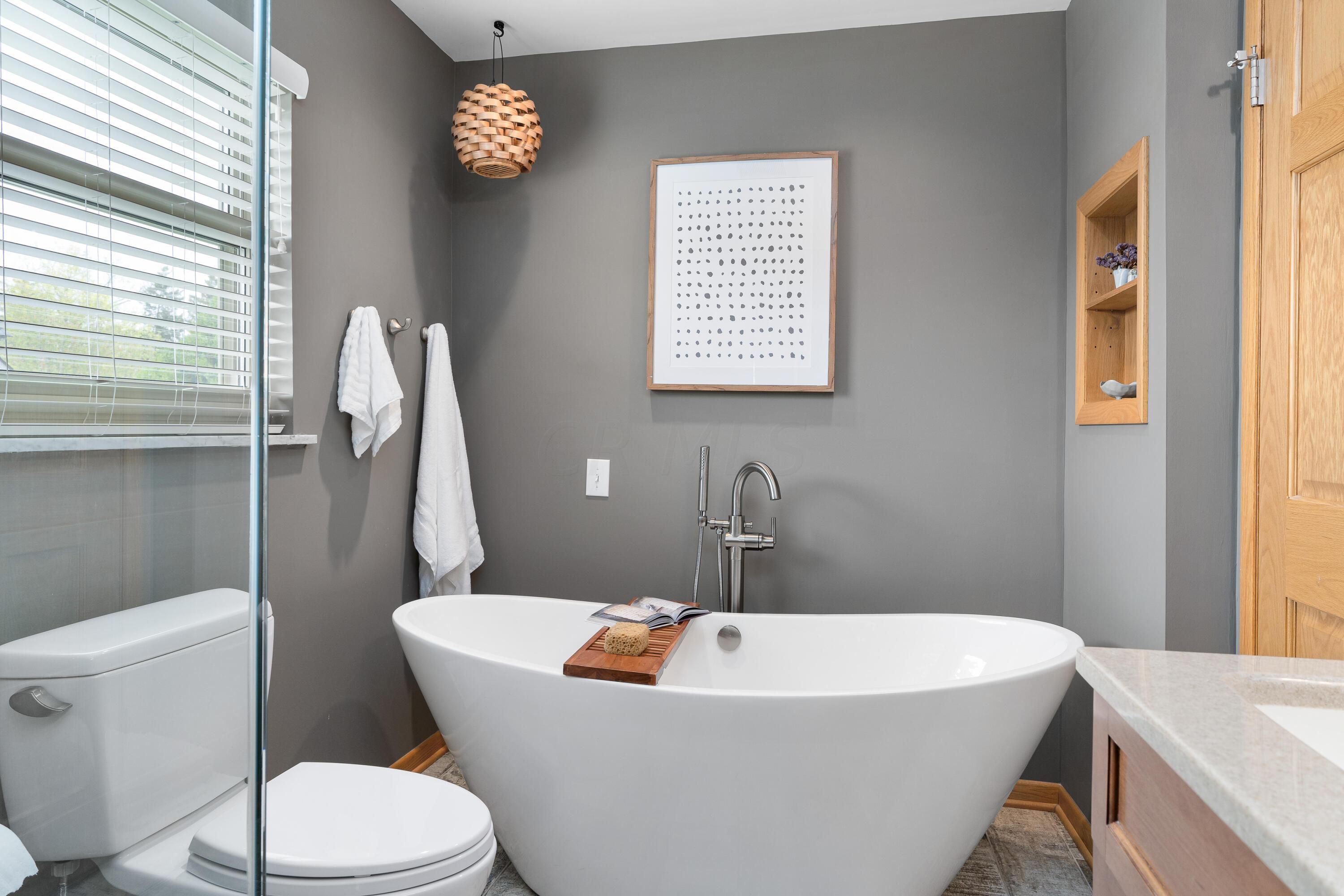 Bathroom with a freestanding soaking tub, glass shower, and gray walls with warm wood accents.