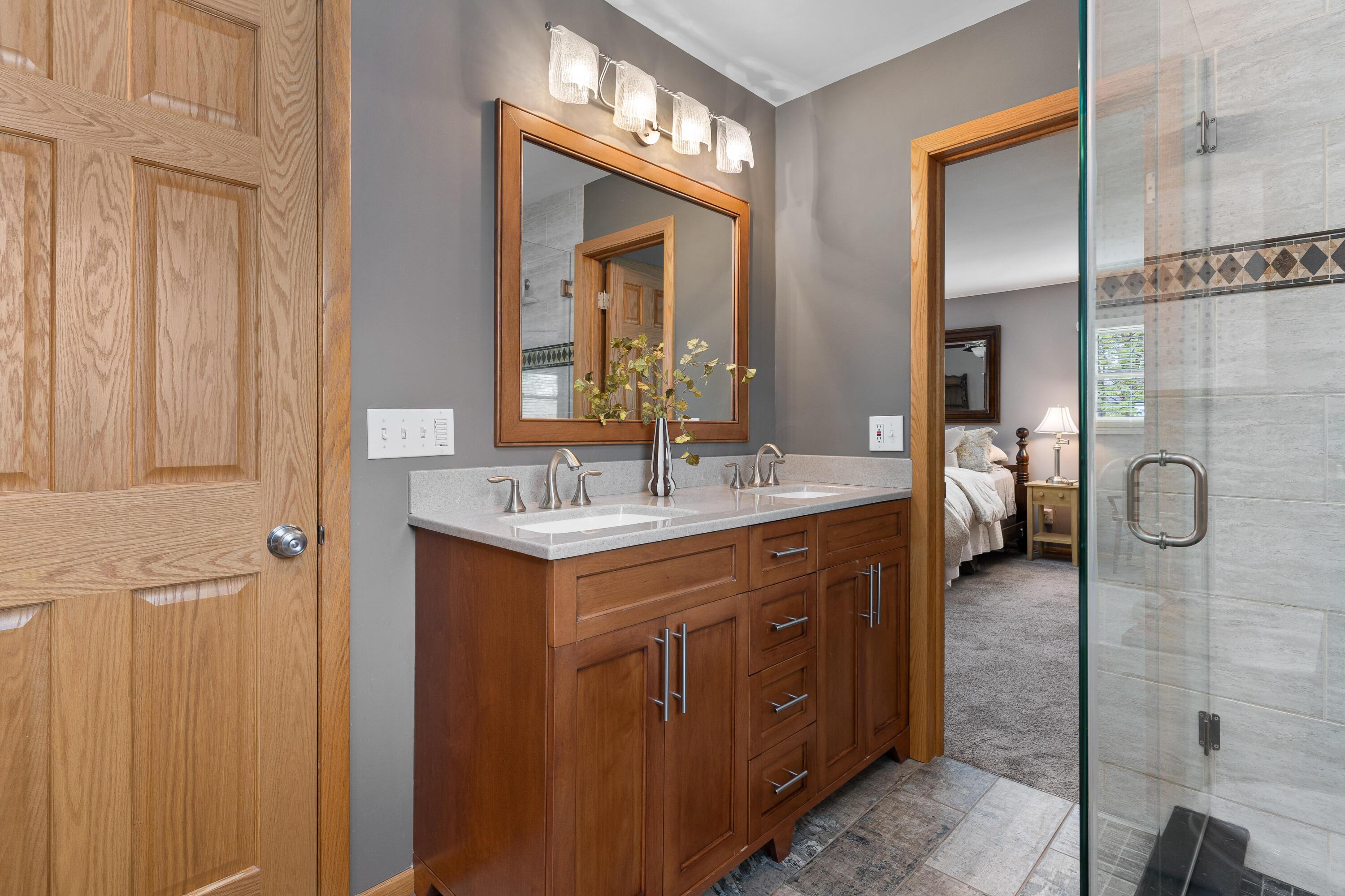Bathroom with a double-sink wood vanity and glass shower, opening onto a bedroom.