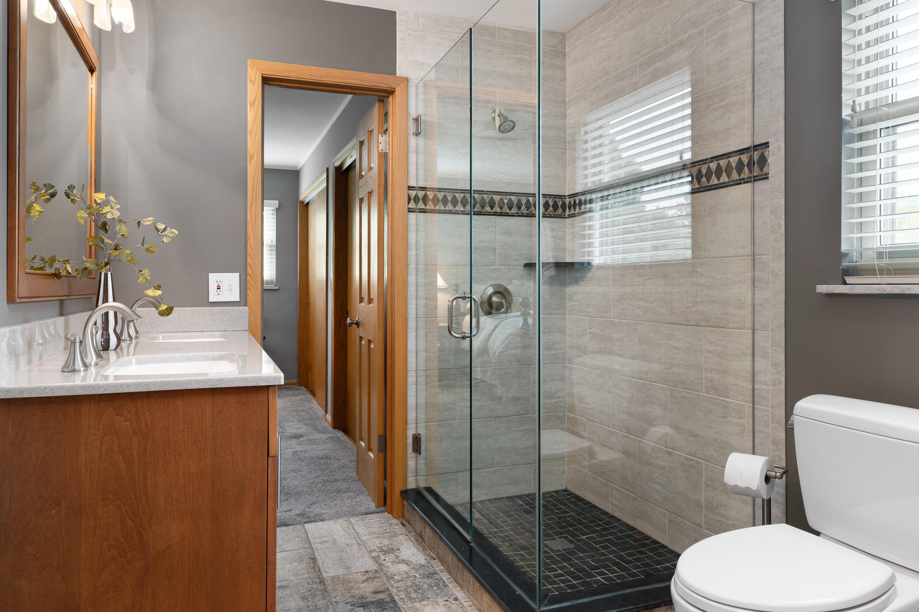 Bathroom with a glass walk-in shower, beige tile, wood vanity, and white toilet.