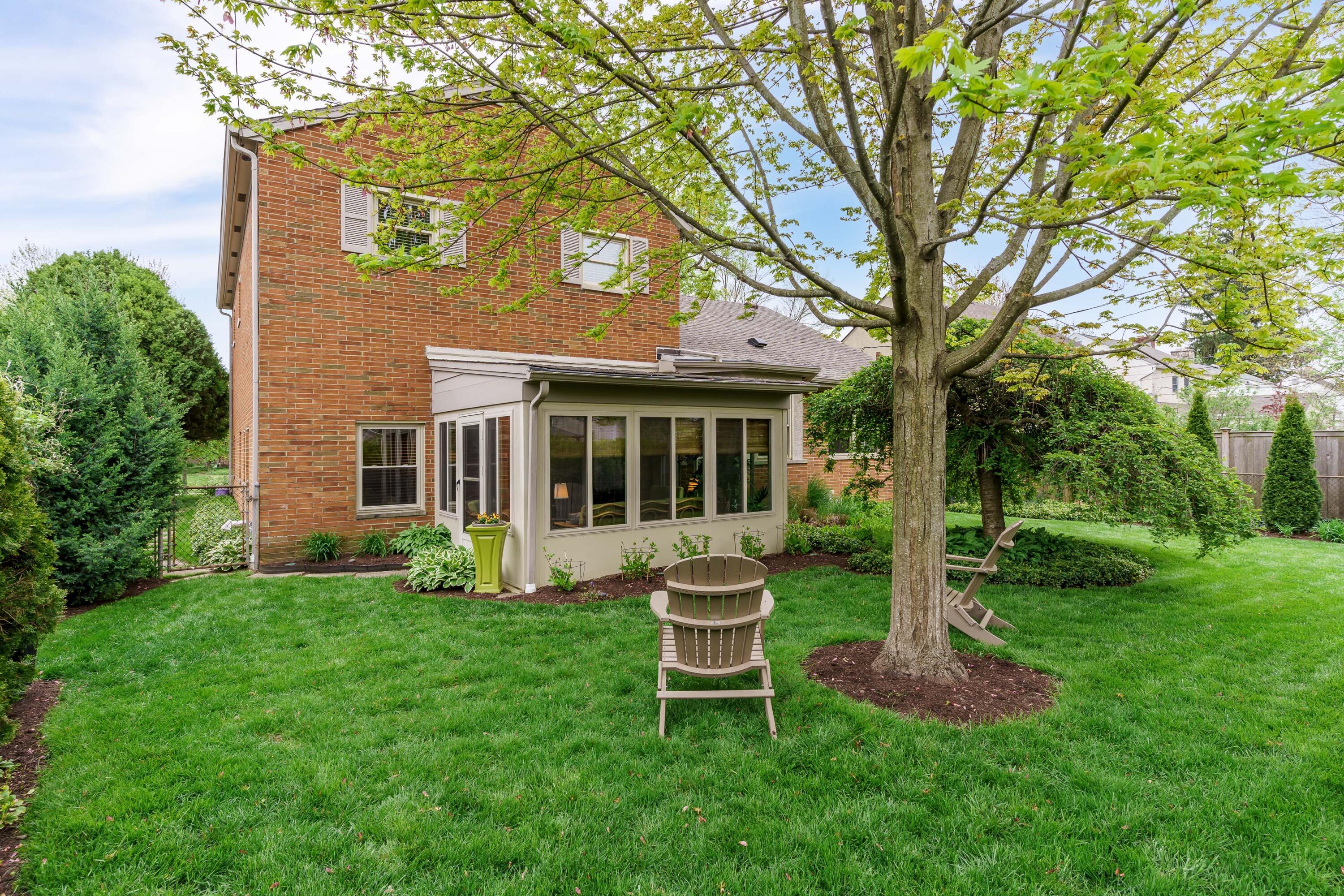 Brick house rear yard with a glass-enclosed sunroom beneath a large tree and Adirondack chairs on the lawn.