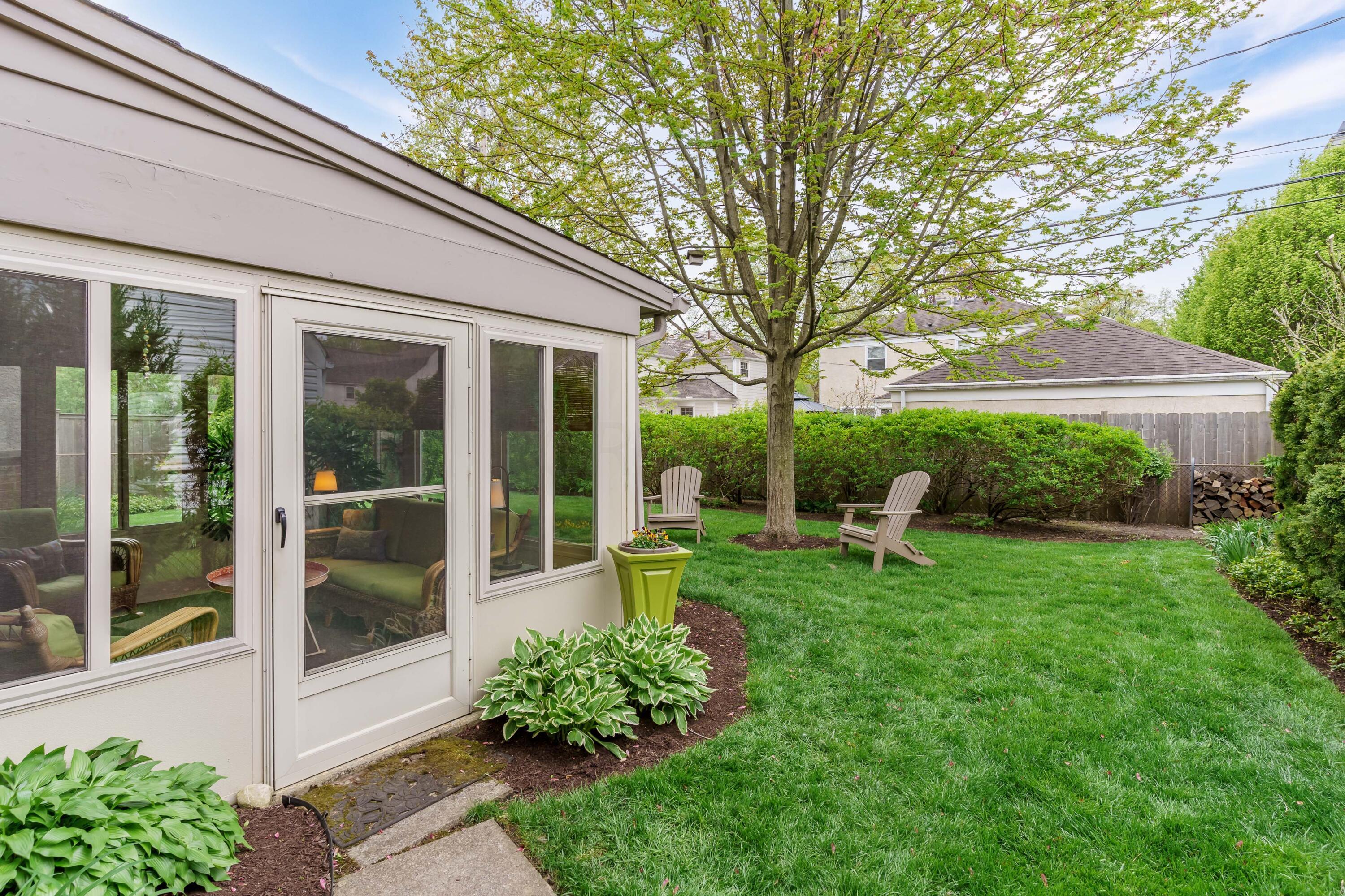Backyard with a white sunroom, green lawn, mature tree, and two Adirondack chairs.