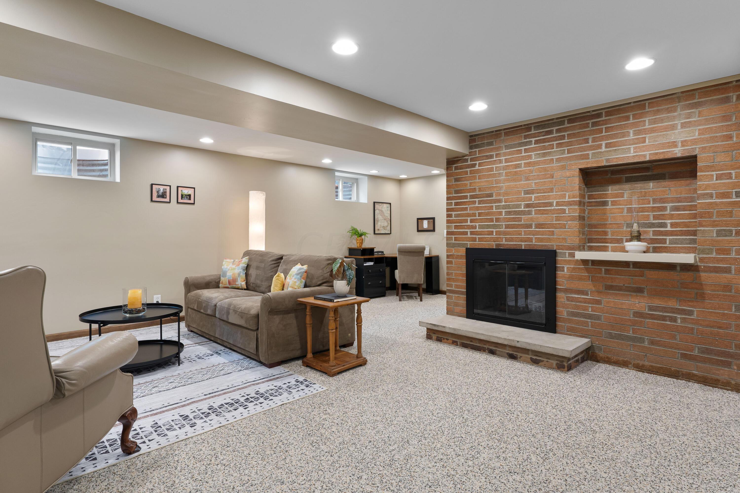 Basement living room with a brick fireplace, gray sofa, and patterned rug.