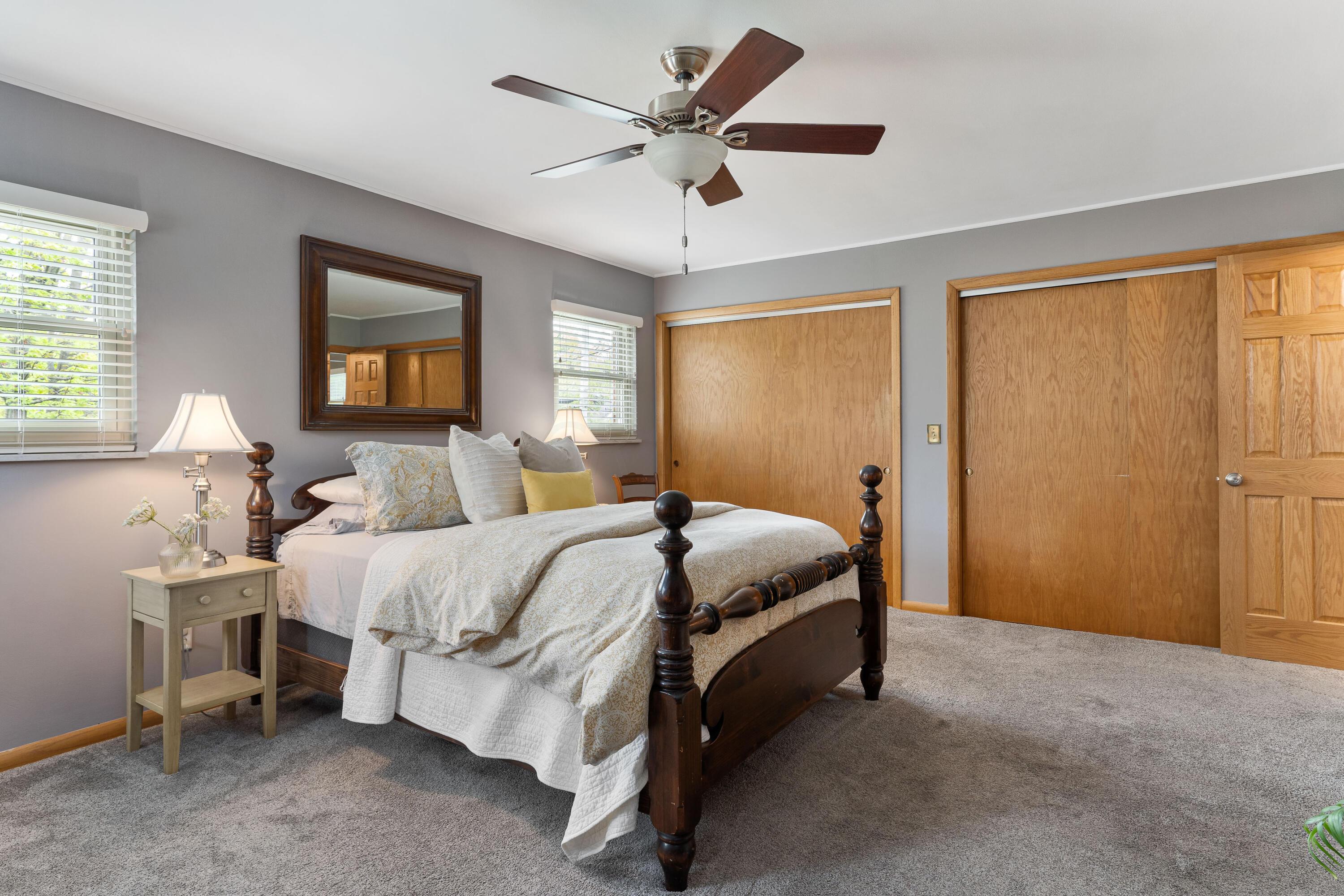 Staged bedroom with a dark wood four-poster bed, gray carpet, ceiling fan, and sliding wood closet doors.