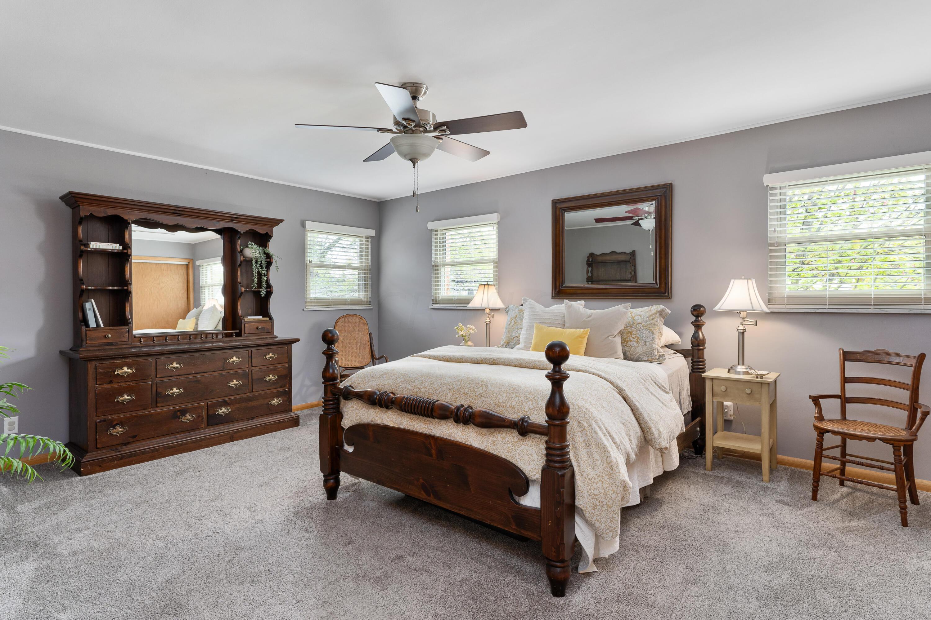 Bedroom with a dark wood four-poster bed, matching dresser, and three windows with white blinds.