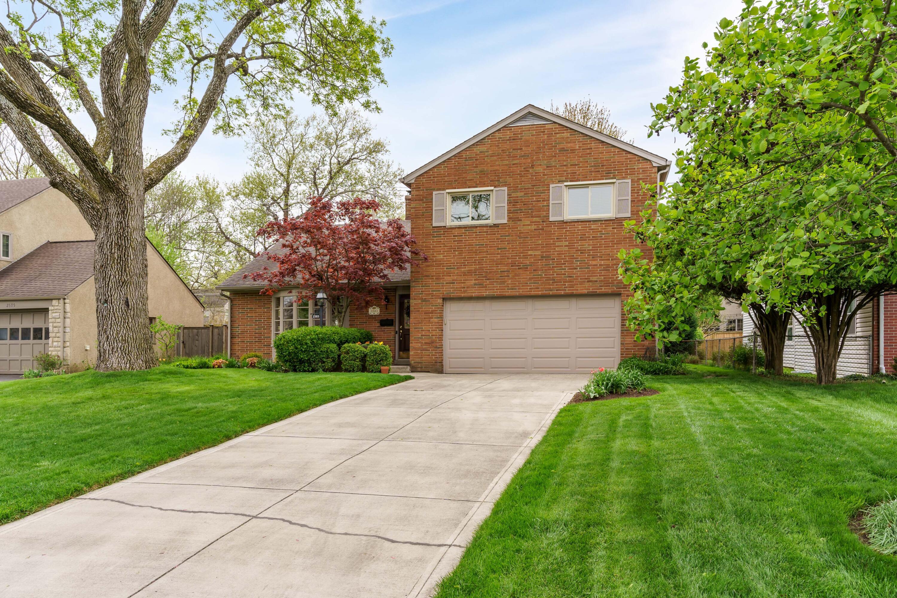 Two-story brick home with an attached garage, wide driveway, and mature trees.