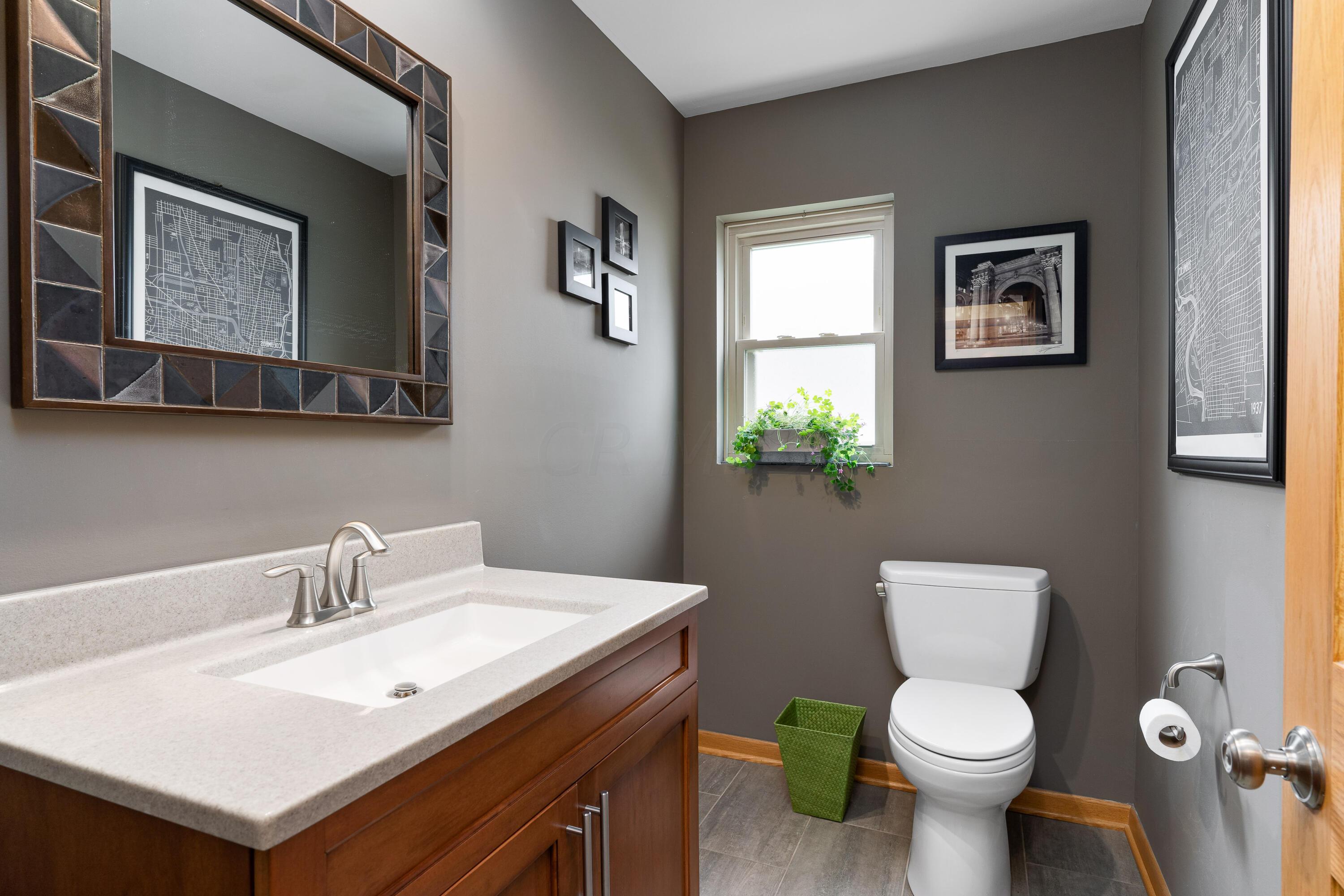 Bathroom with a wood vanity, pale countertop, white toilet, and a small window with a potted plant.