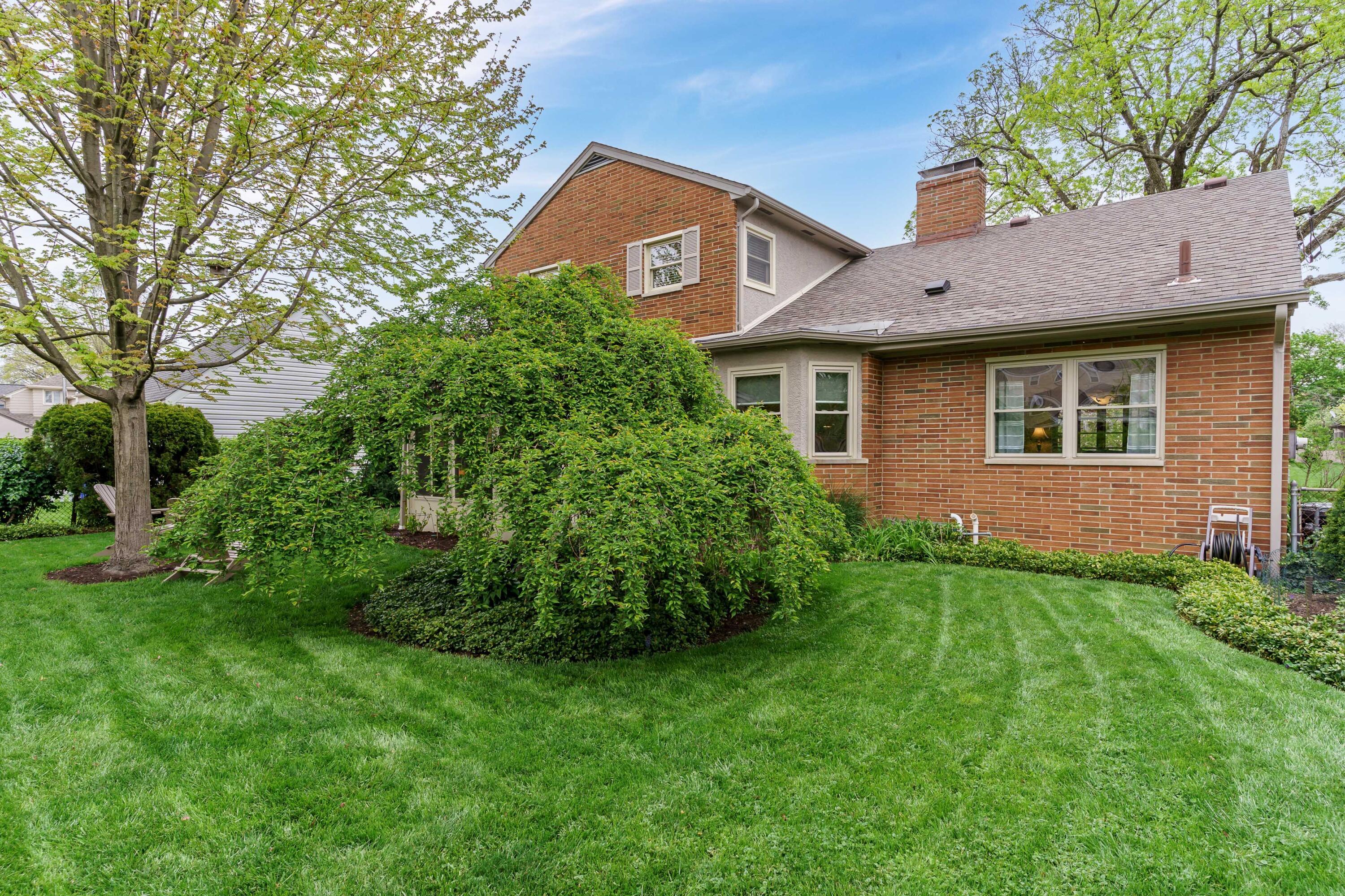 Two-story brick house viewed from the landscaped backyard, with mature trees and a broad green lawn.