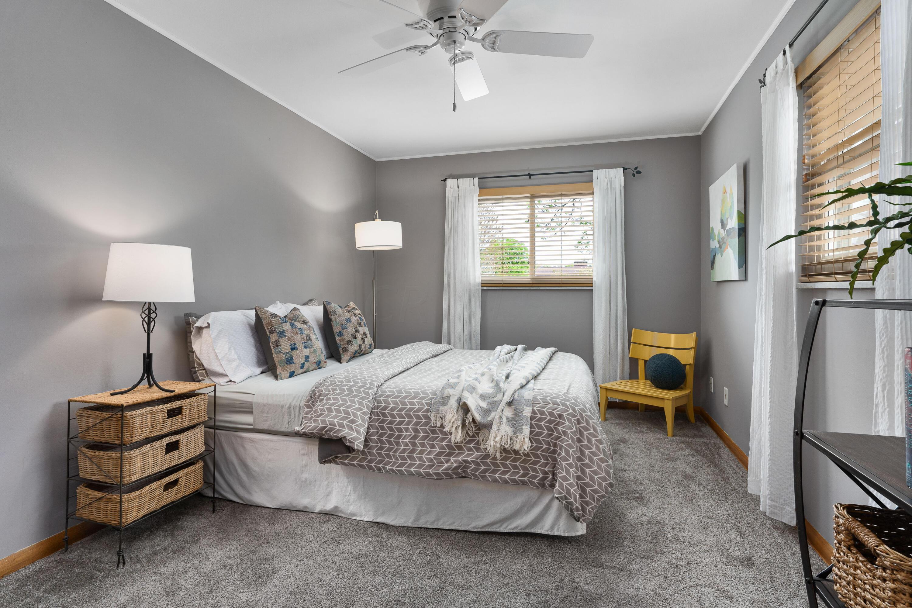 Furnished bedroom with gray patterned bedding, two curtained windows, and a mustard-yellow chair.