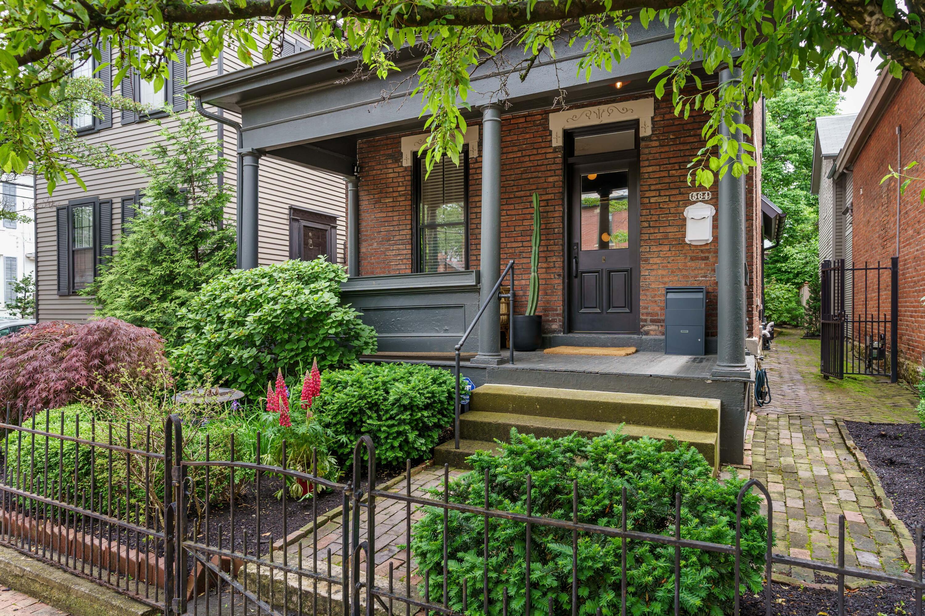 Brick home with a covered porch, lush front garden, iron fence, and brick walkway.