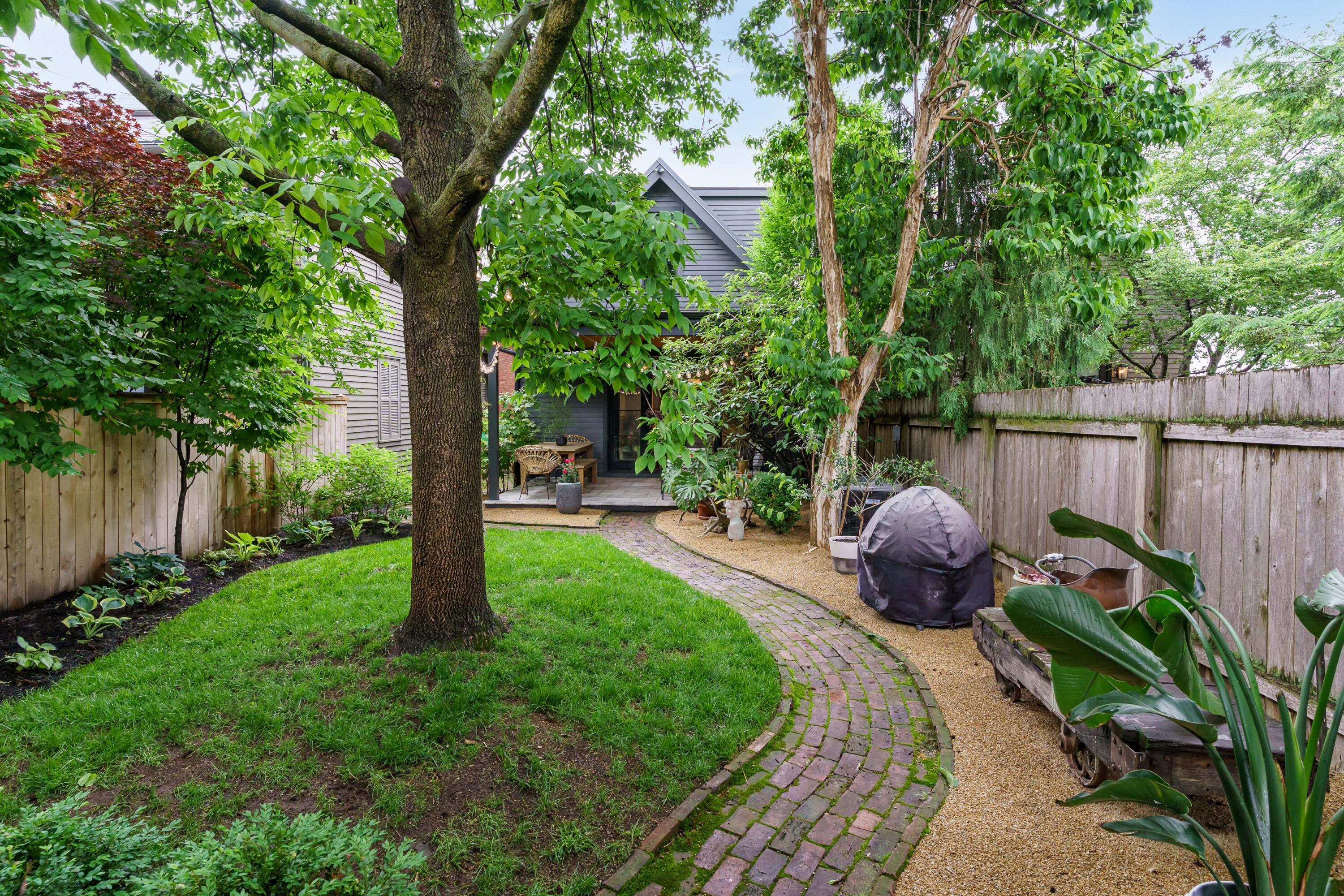 Leafy backyard with a curved brick path, mature shade tree, garden beds, and a small patio.