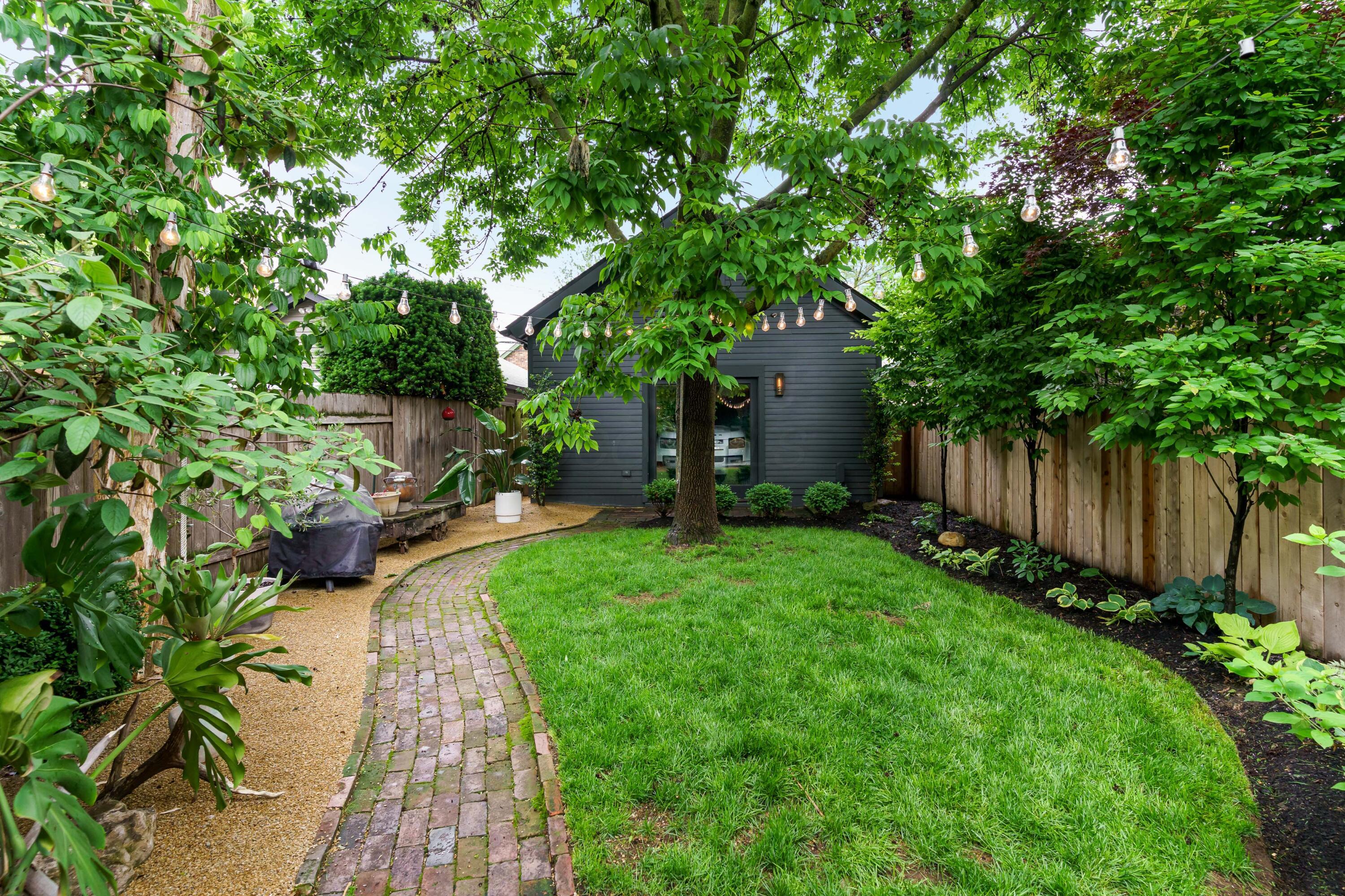 Fenced backyard with a curved brick path, mature trees, lawn, and string lights.
