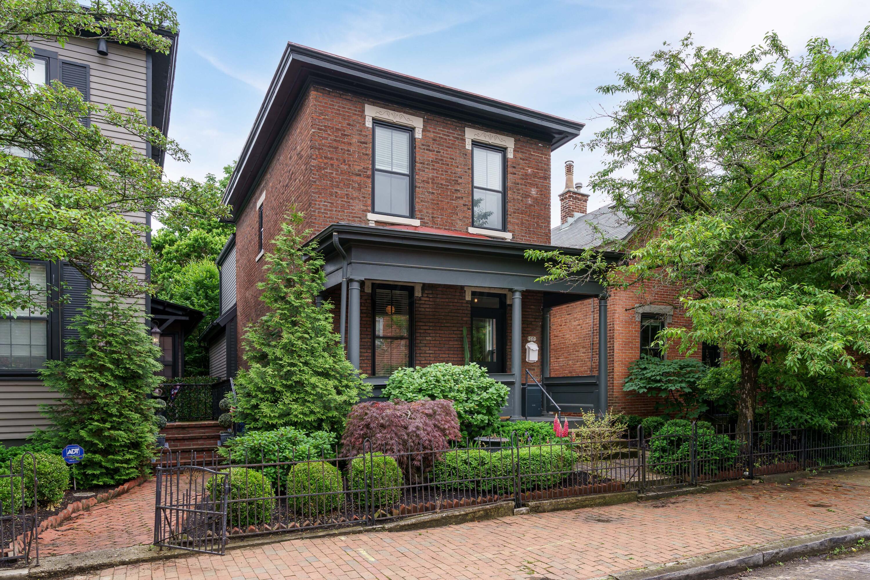 Two-story red-brick house with a covered porch, leafy garden, and black iron fence.