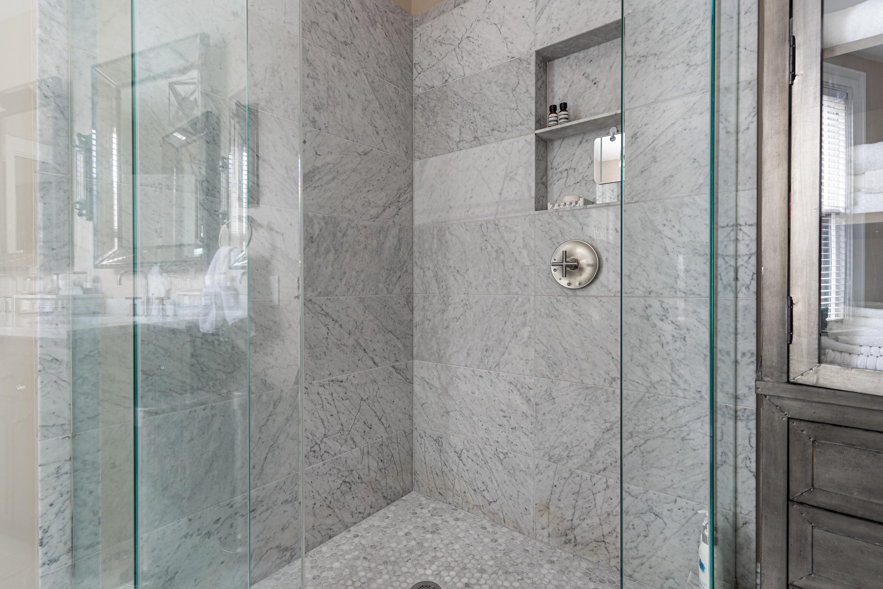 Marble-tiled walk-in shower with glass enclosure, recessed shelf, and hexagonal mosaic floor.