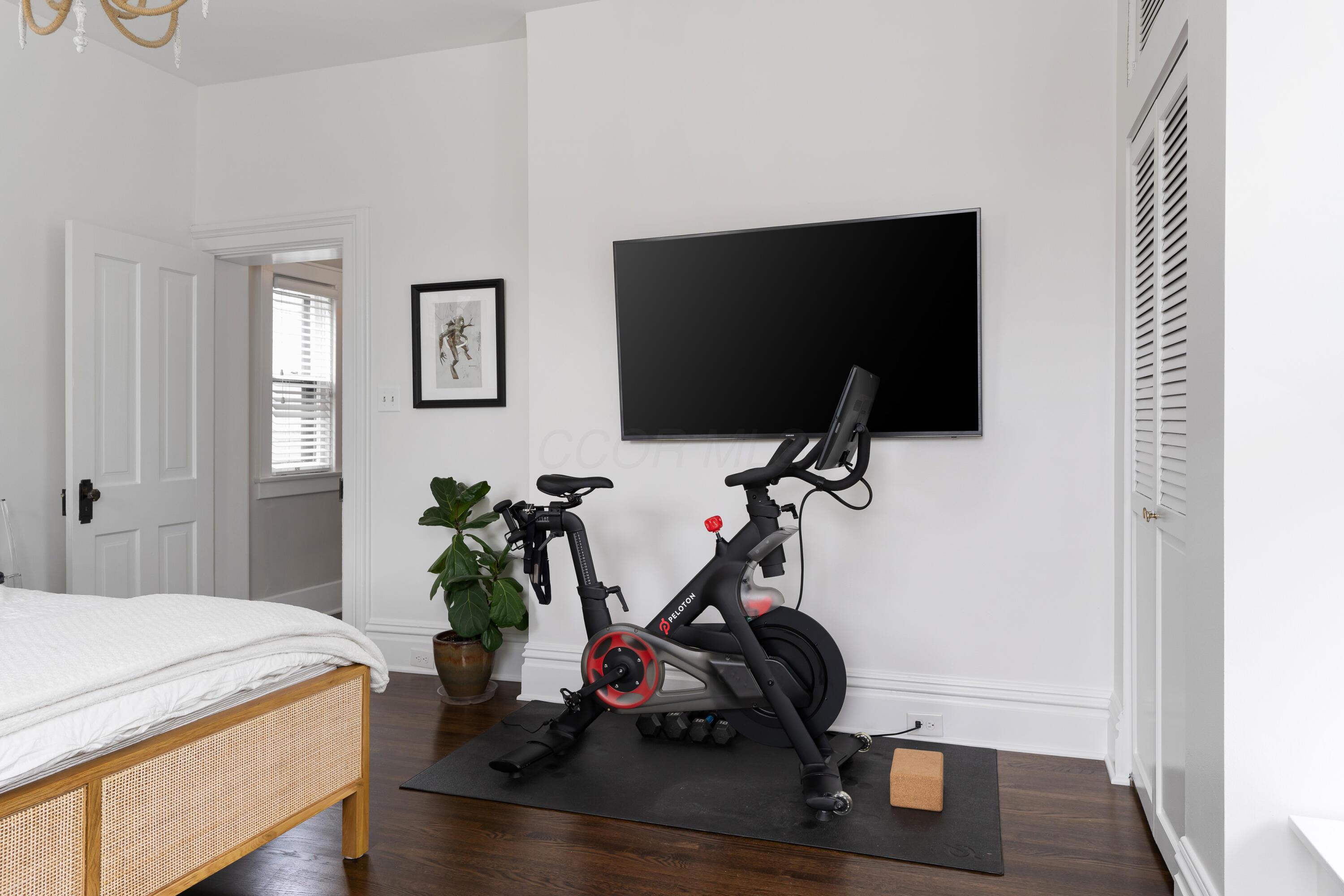 Bedroom with a Peloton bike and weights beside a woven cane bed.