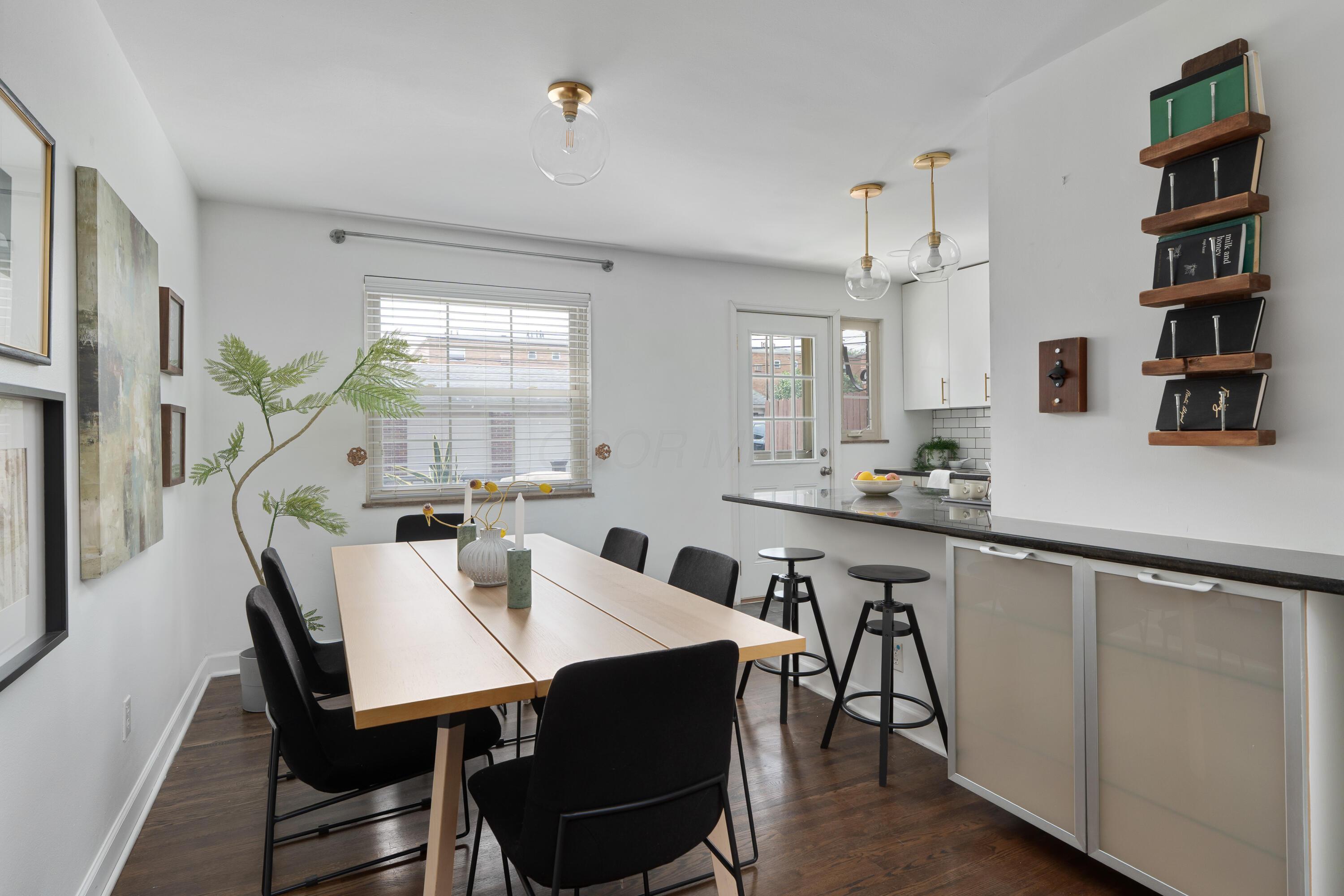Dining area with a light wood table and black chairs beside a kitchen breakfast bar and two stools.
