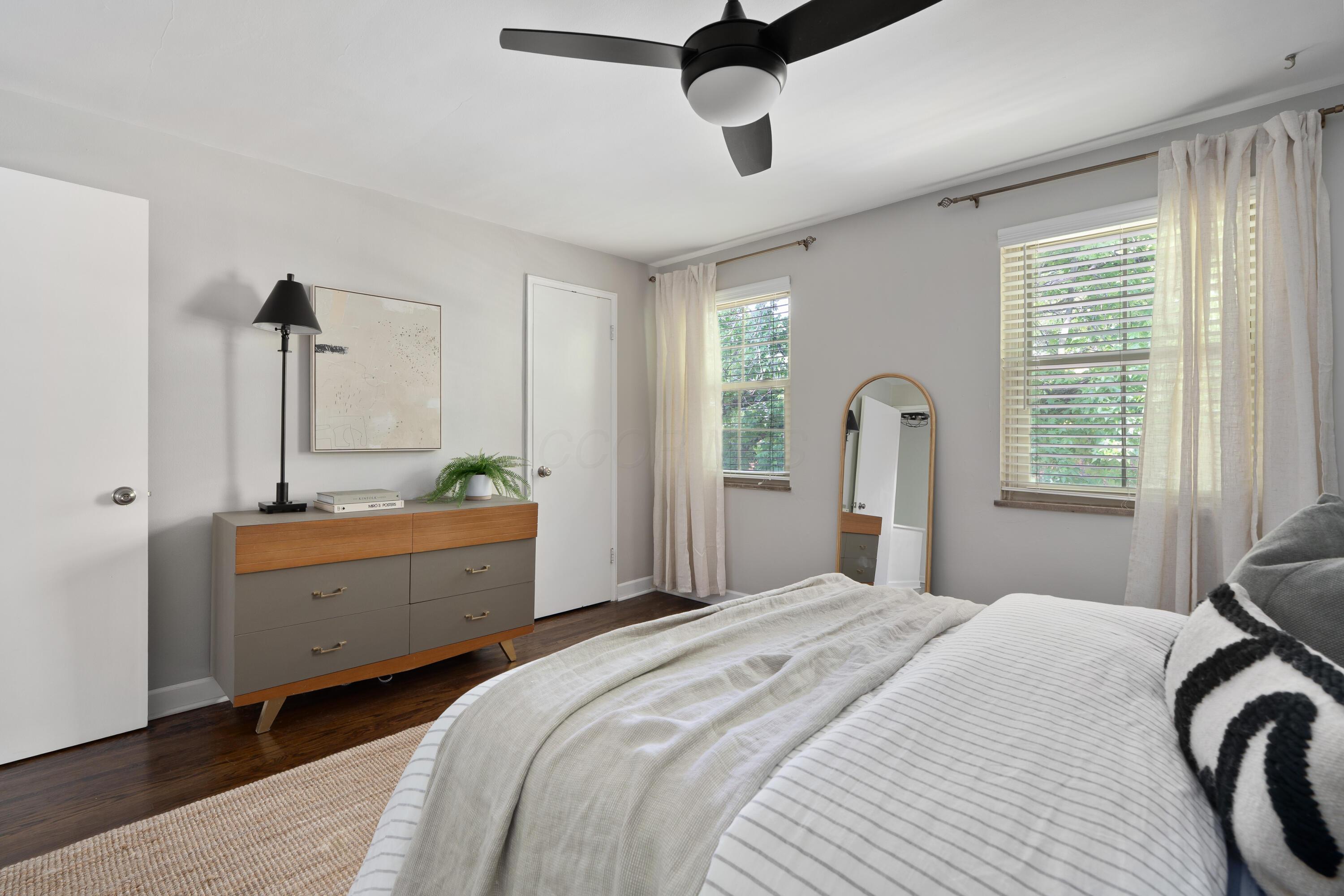 Furnished bedroom with a striped bed, modern dresser, standing mirror, and two curtained windows.