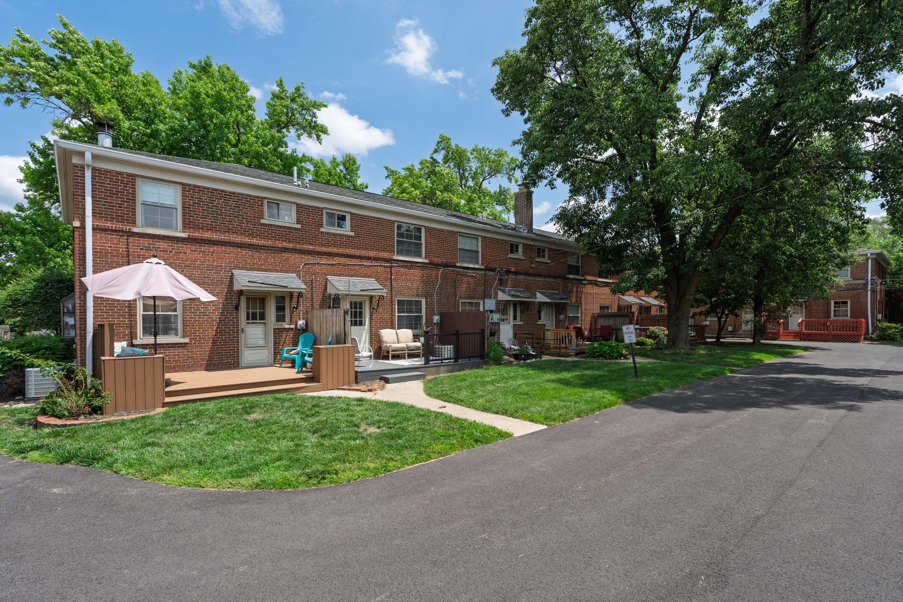 Two-story brick residential building with individual patios, outdoor seating, and a large shade tree.