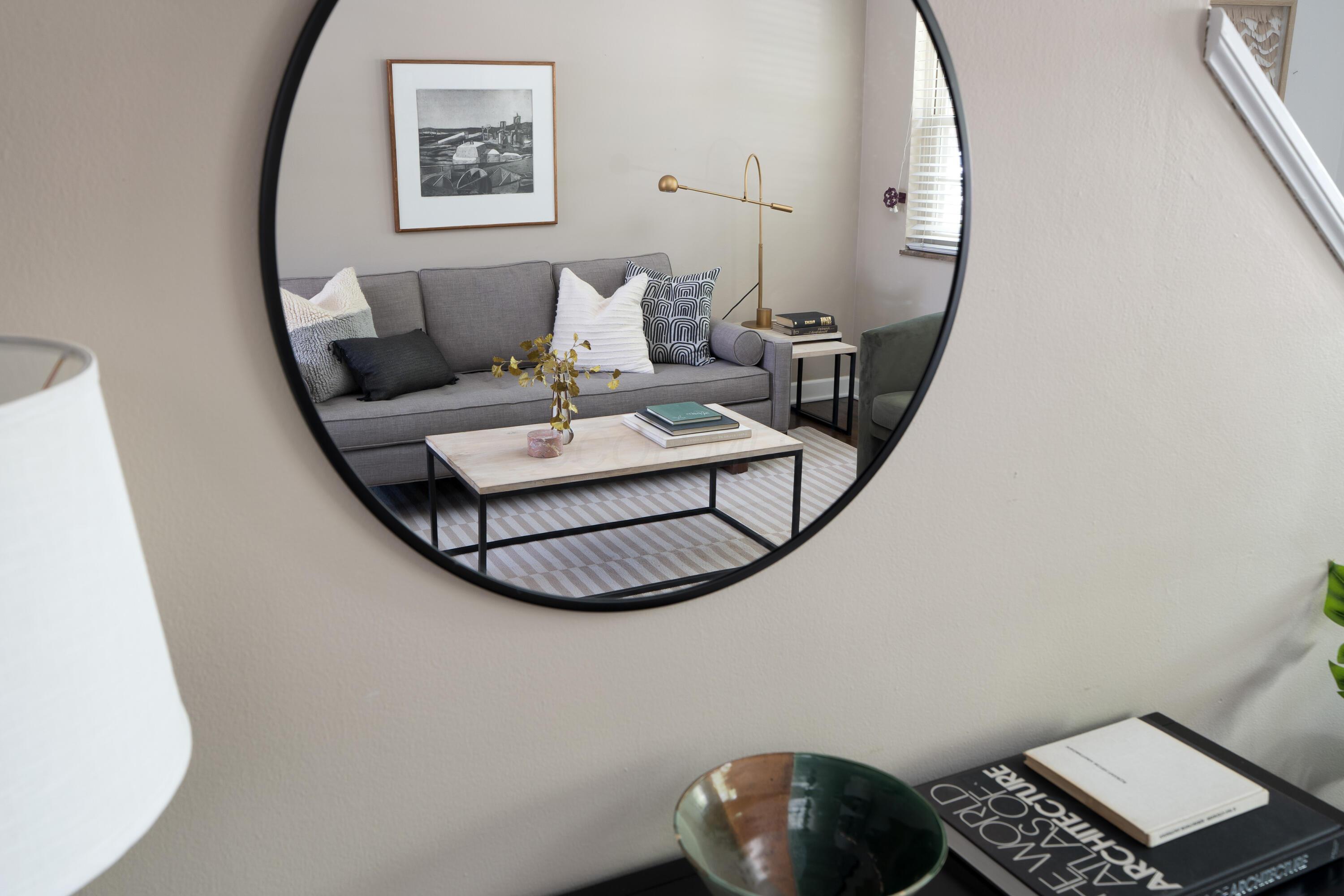 Staged living room reflected in a round mirror, with a gray sofa, brass lamp, and striped rug.