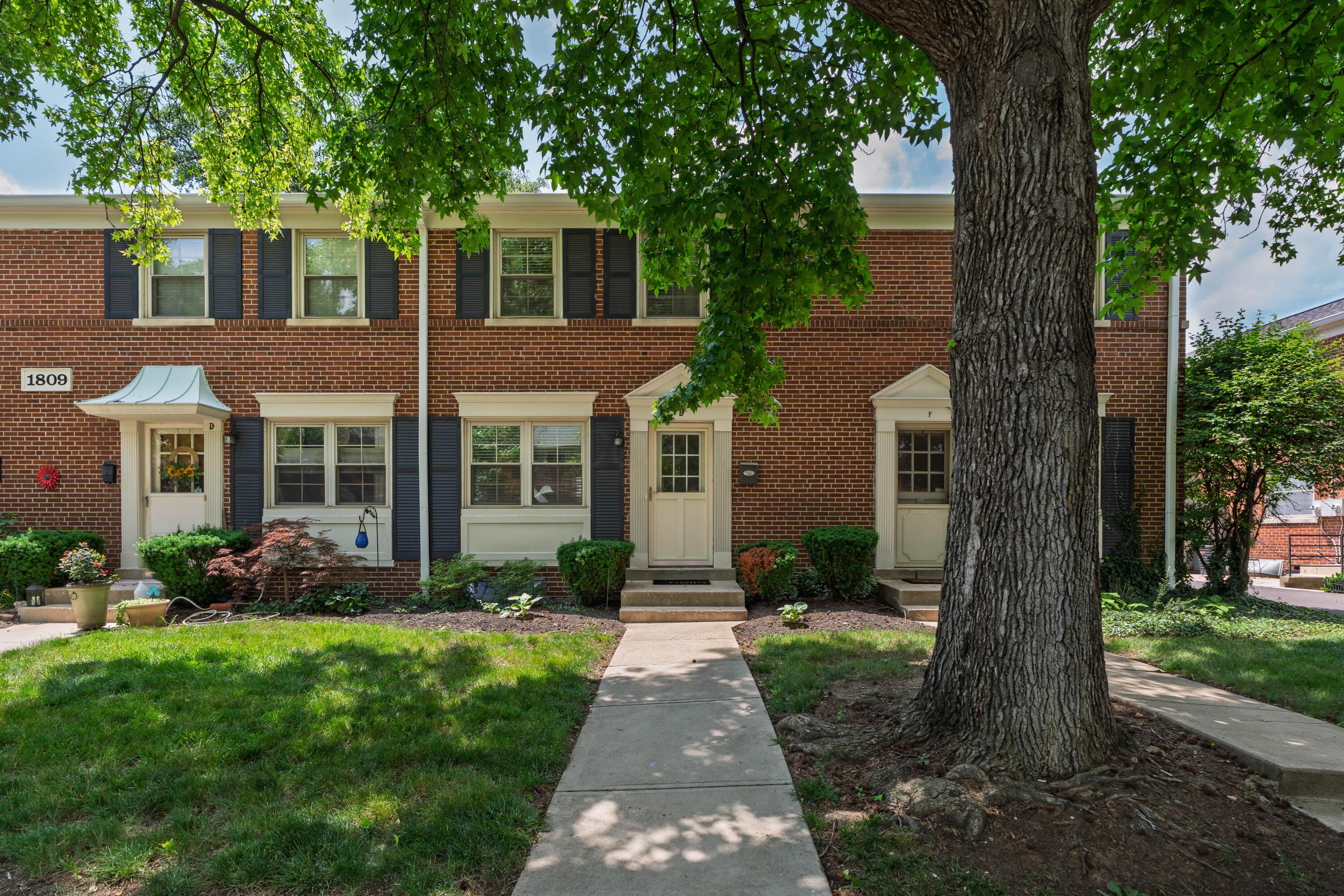 Red-brick attached residence with cream-trimmed entrances, landscaped lawn, and a large shade tree.
