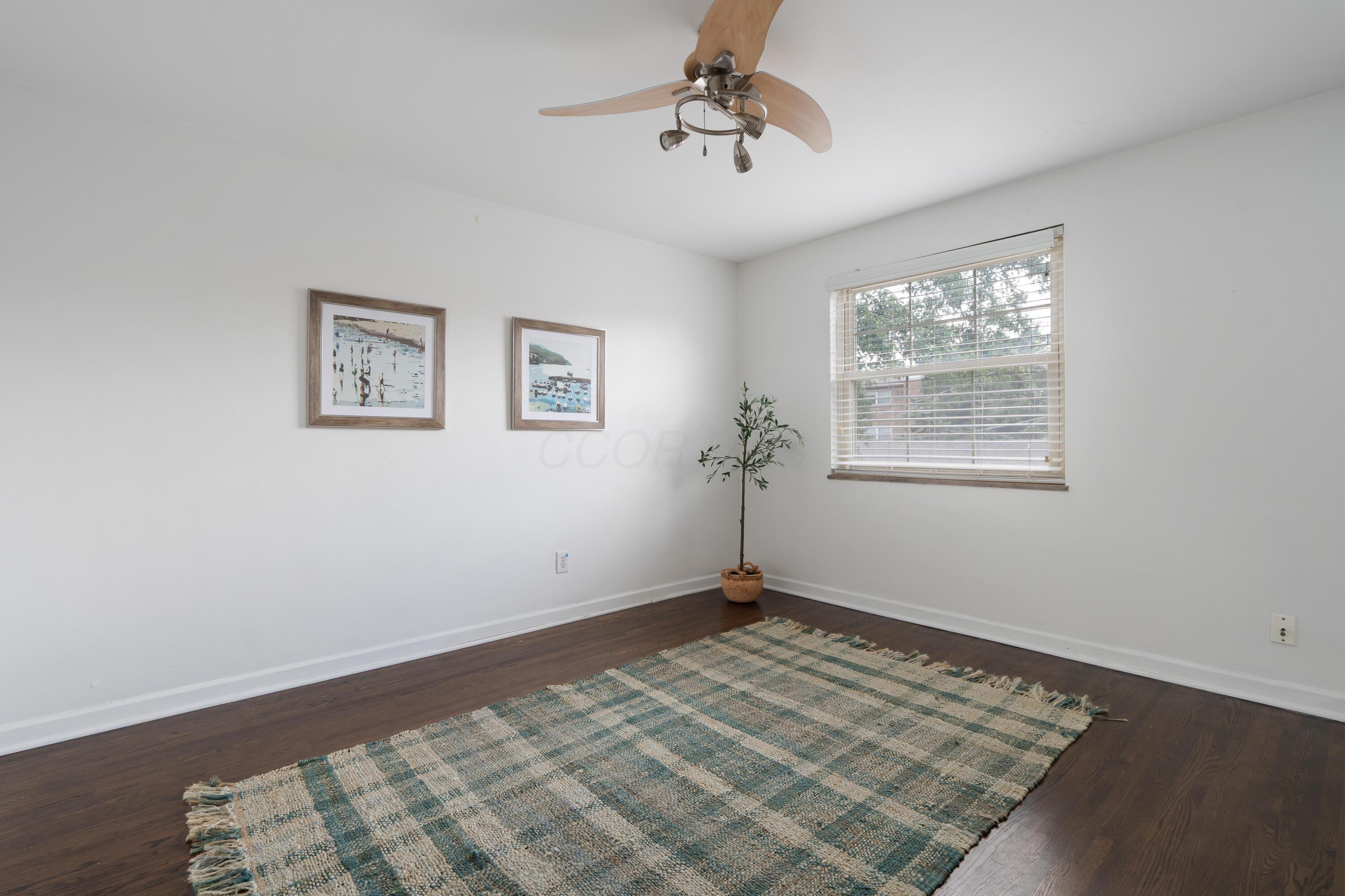 Bright room with dark hardwood floors, a blue-and-cream rug, ceiling fan, and window with blinds.