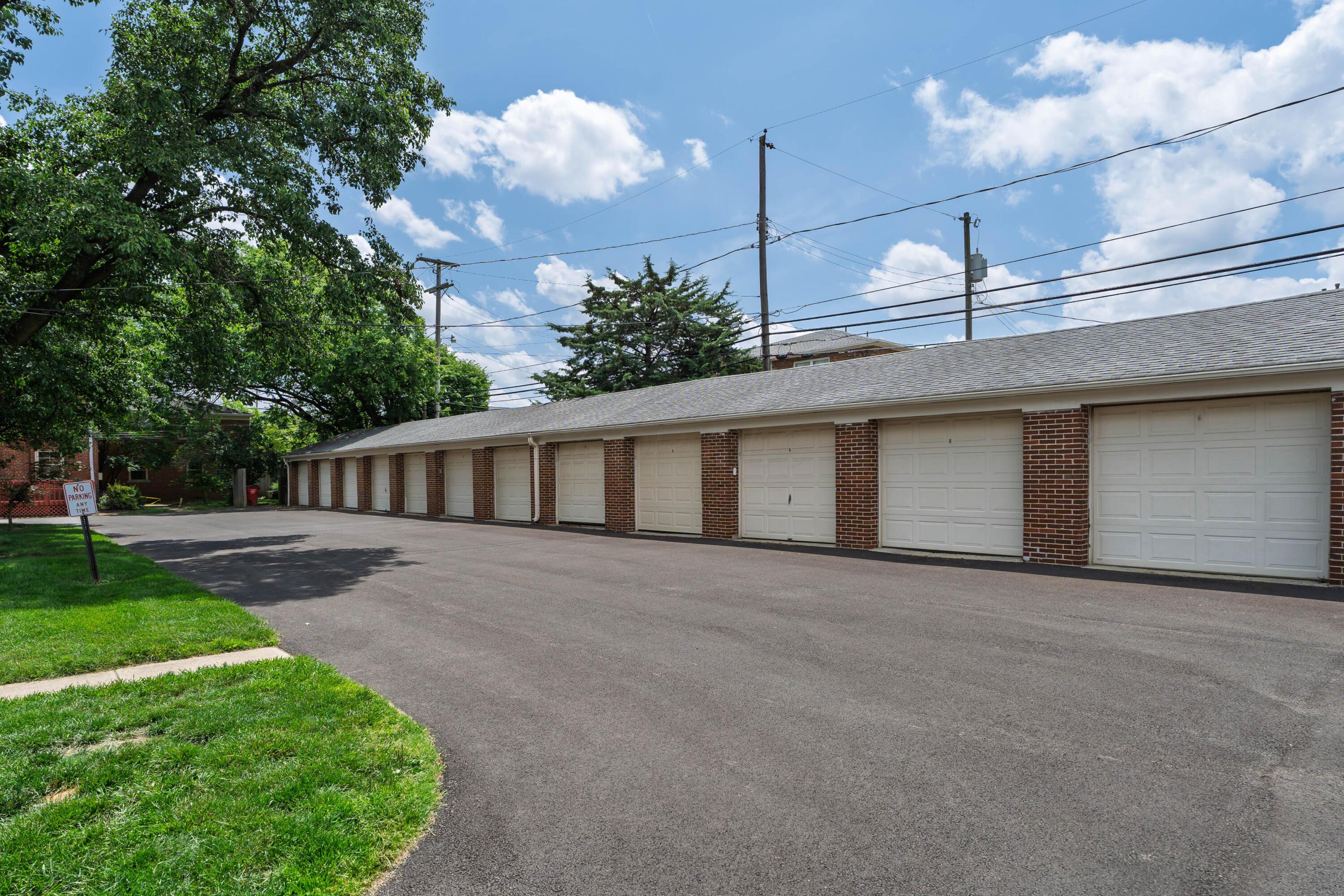 Row of white garage doors in a brick structure beside a paved driveway and mature trees.