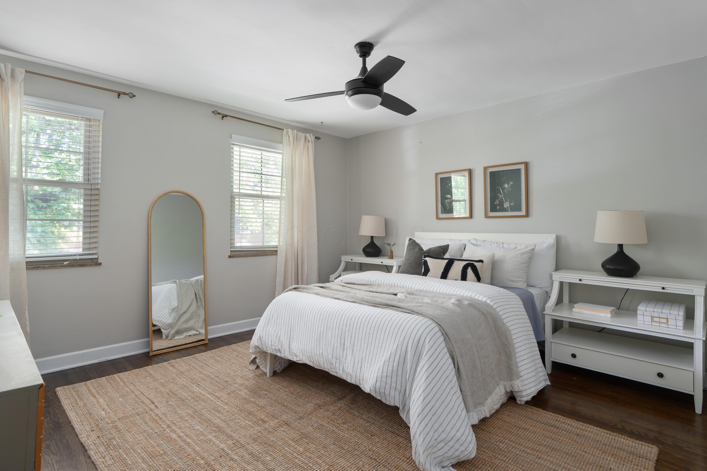 Furnished bedroom with a striped bed, arched mirror, two windows, and a black ceiling fan.