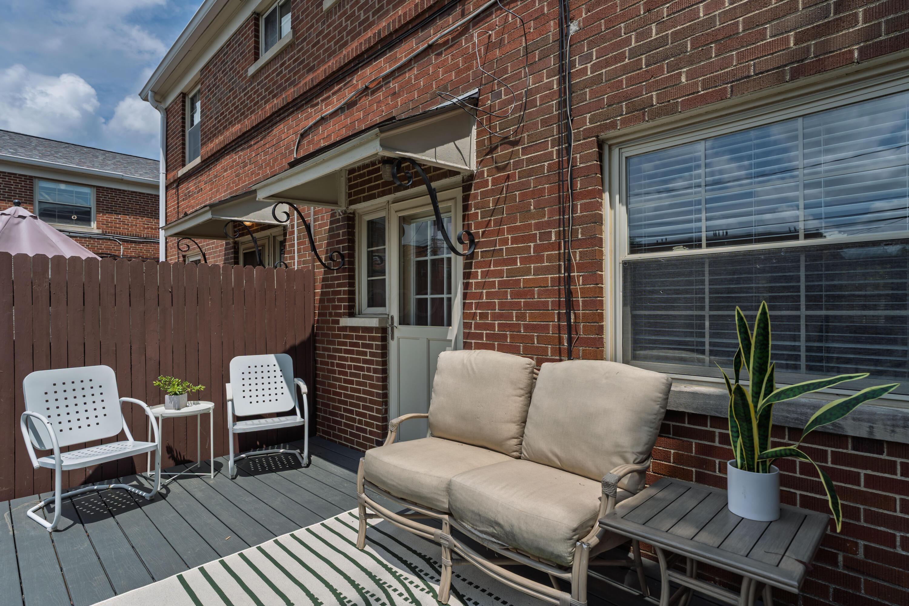 Furnished deck beside a red-brick home, with a cushioned loveseat and white chairs.