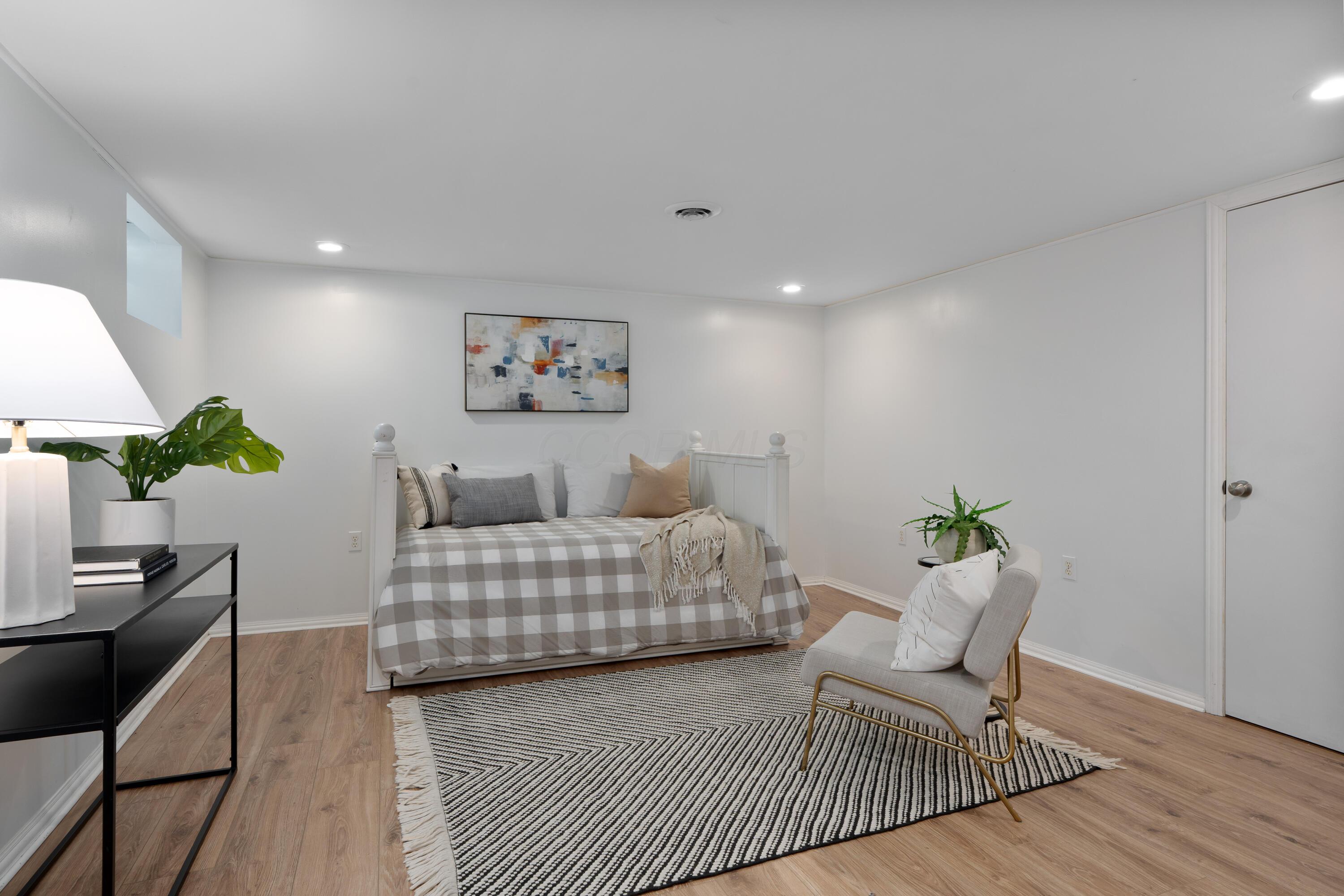 Staged bedroom with a white daybed, gray check bedding, patterned rug, and wood-look floors.