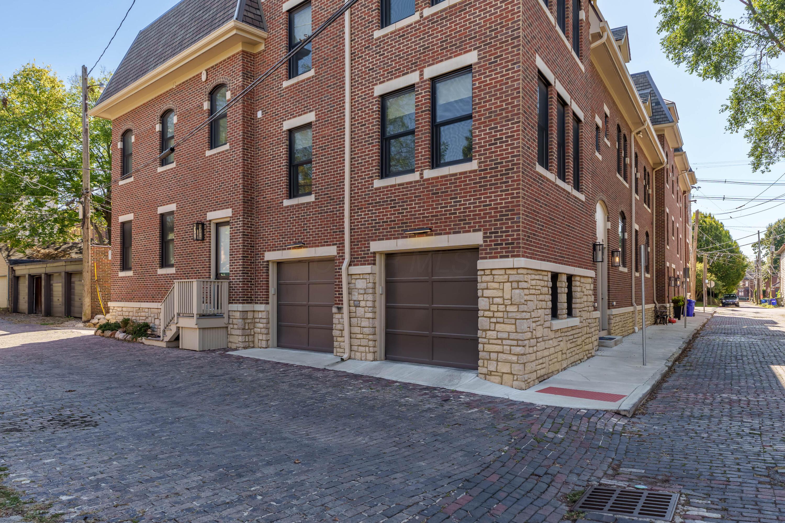 Red-brick building with stone base and two dark garage doors beside a cobblestone street.