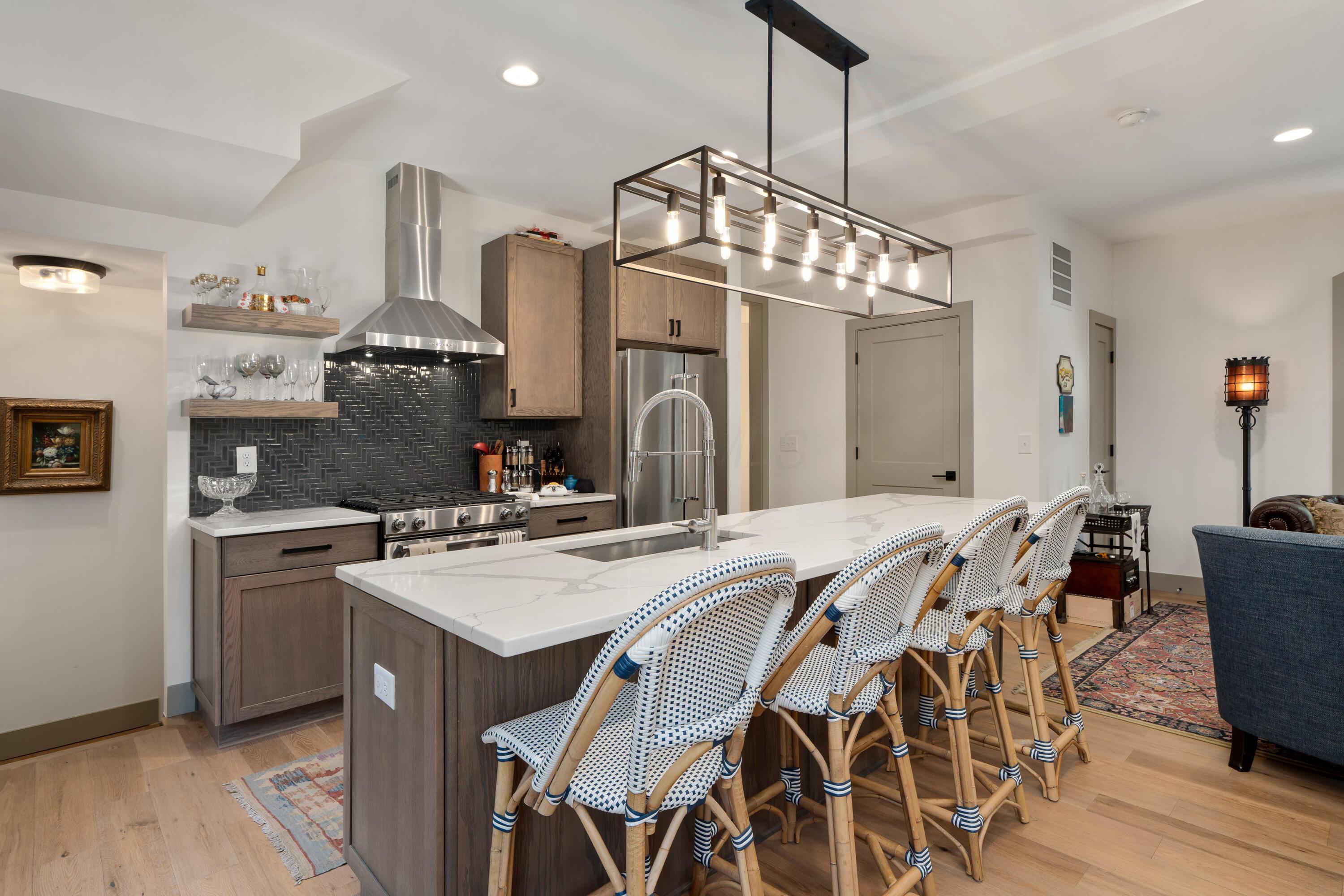Open-plan kitchen with a marble-look island, woven stools, dark backsplash, and stainless steel appliances.