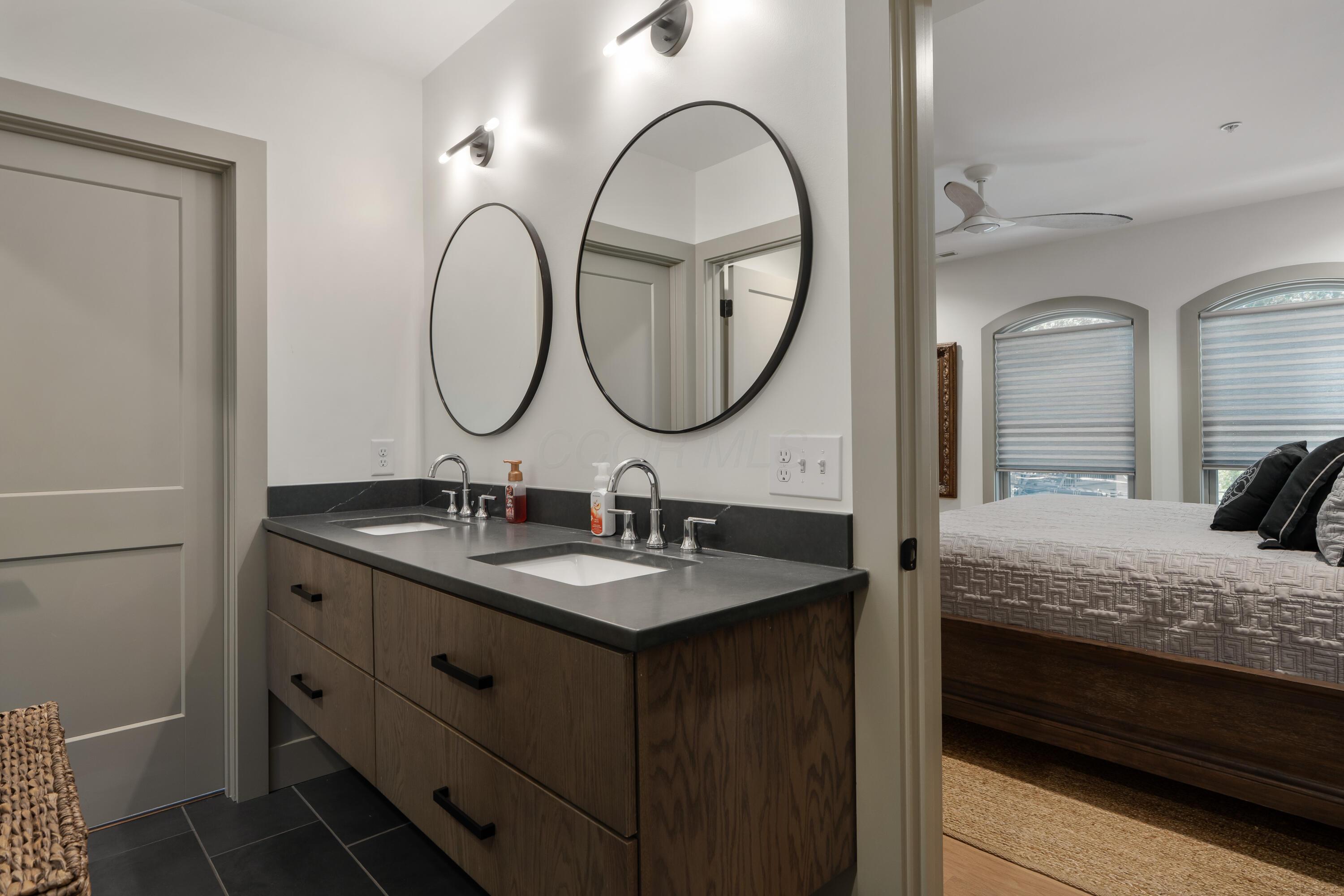 Bathroom with a dark wood double vanity and round mirrors, opening to a bedroom with arched windows.