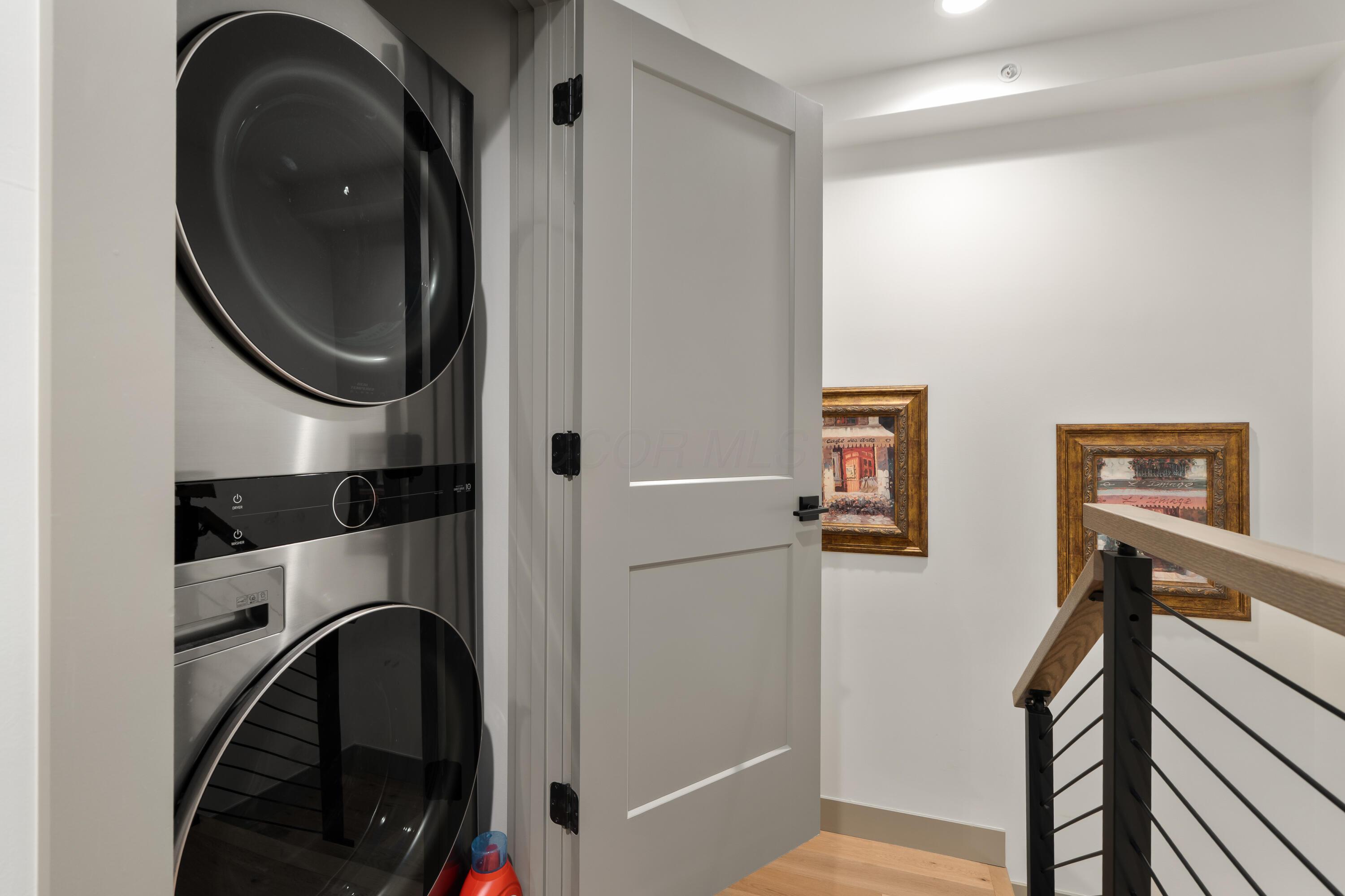 Stacked stainless-steel washer and dryer beside a stair landing with framed artwork.