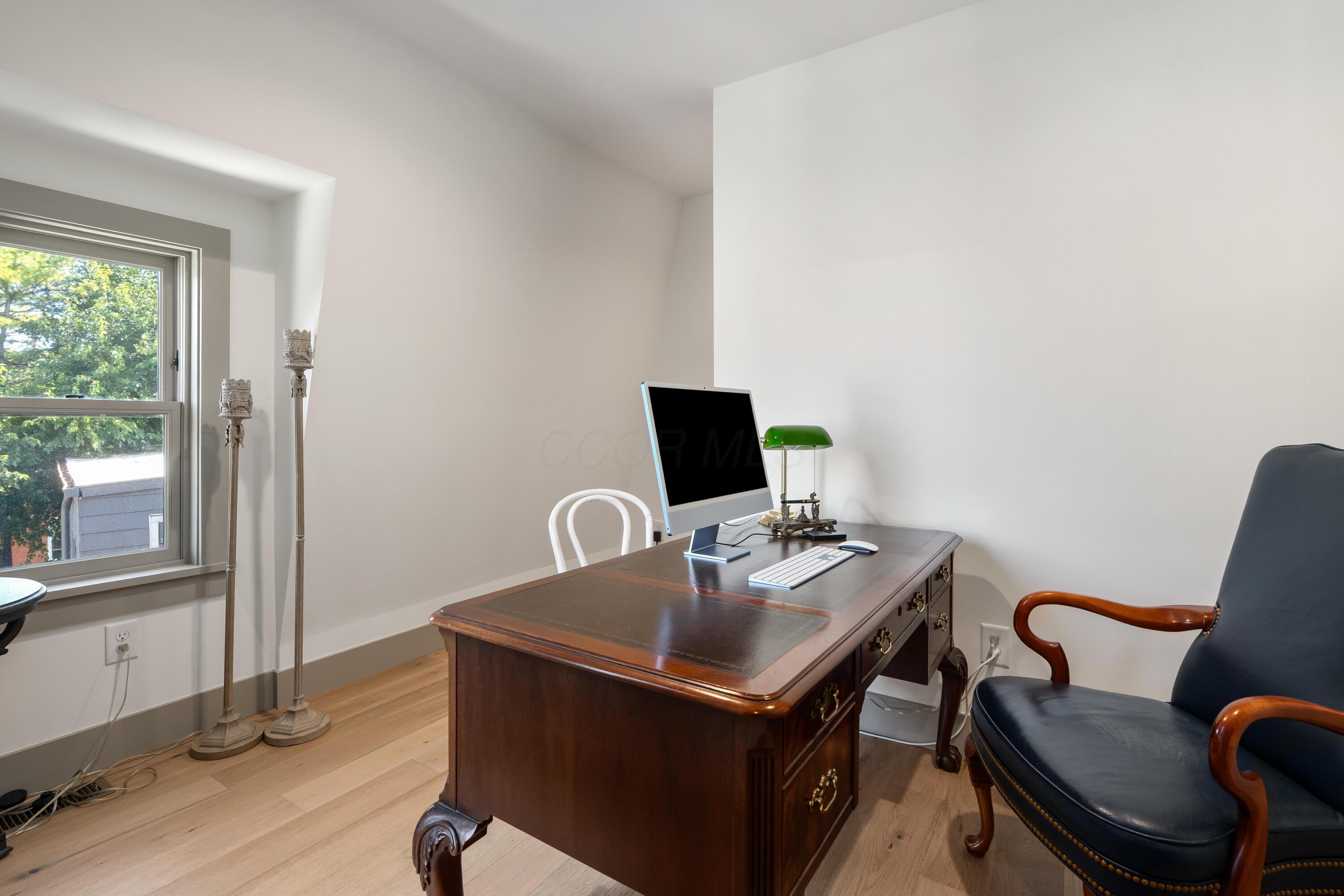 Furnished home office with a carved wooden desk, leather armchair, and window overlooking leafy trees.
