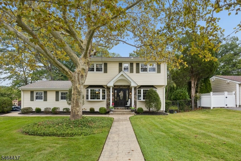 Two-story cream-sided home with a black front door, mature trees, and a paved walkway across the lawn.
