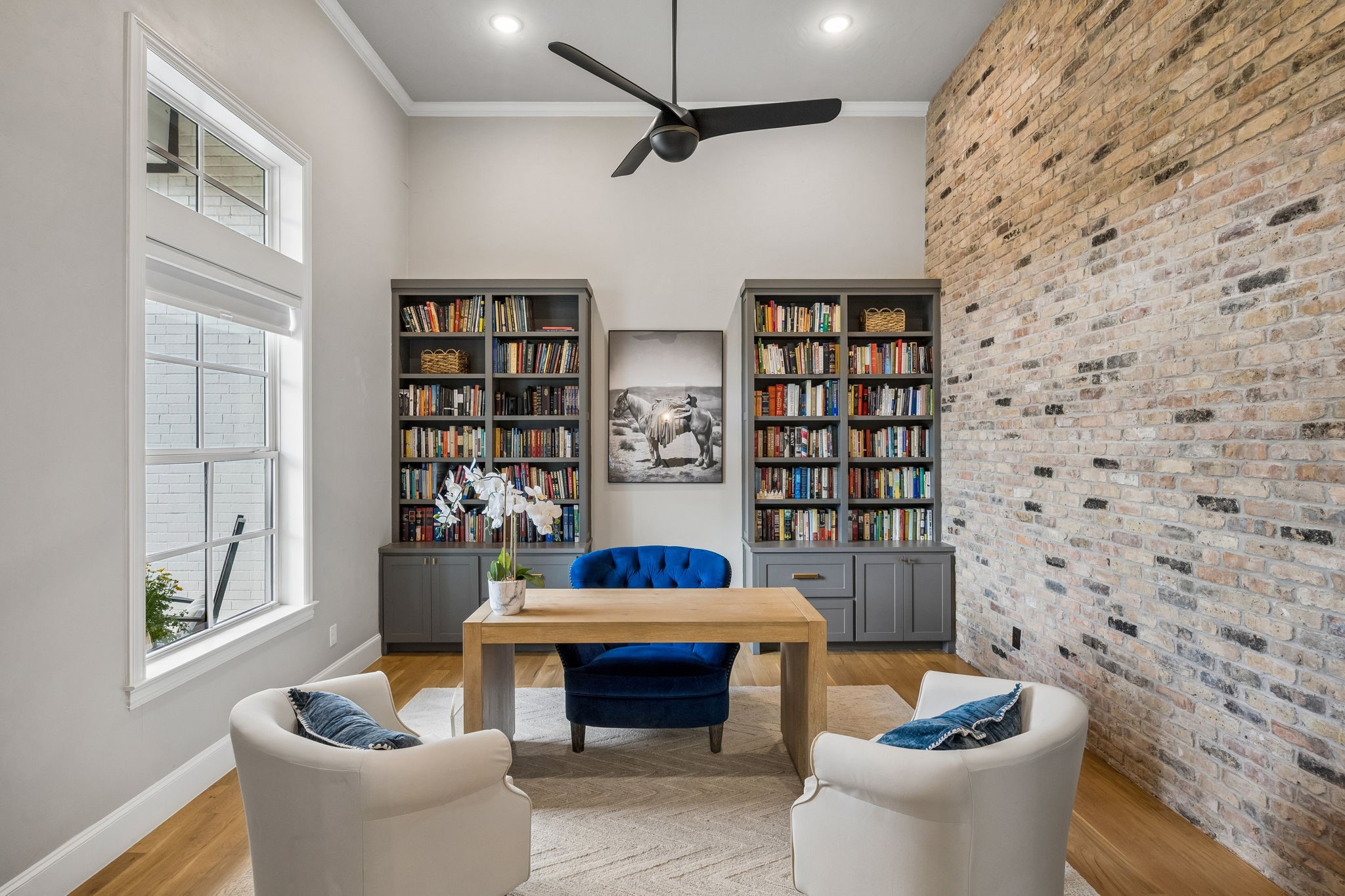 Home office with book-filled built-ins, a wooden desk, blue chair, and exposed-brick wall.