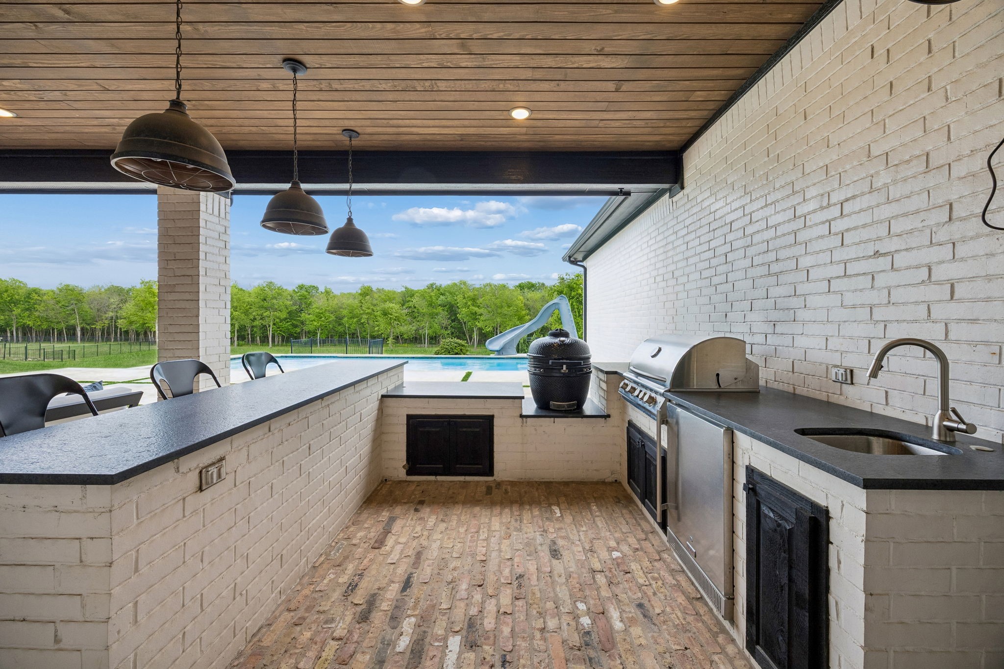 Covered outdoor kitchen with brick counters and stainless grill overlooking a pool and wooded yard.