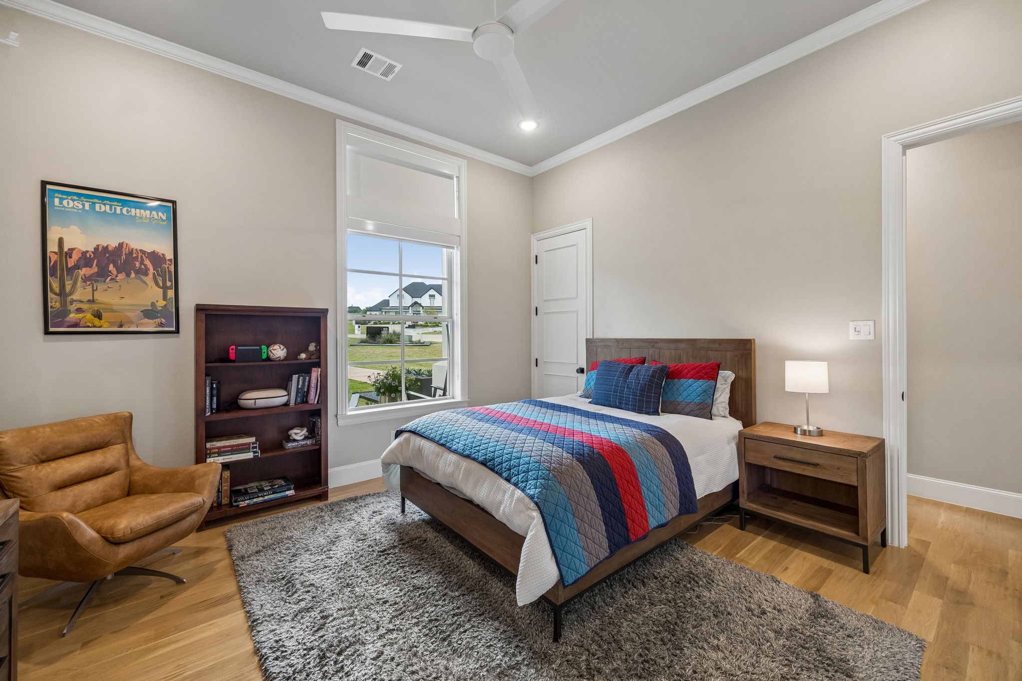 Furnished bedroom with a blue-and-red quilted bed, bookcase, and leather chair beside a large window.