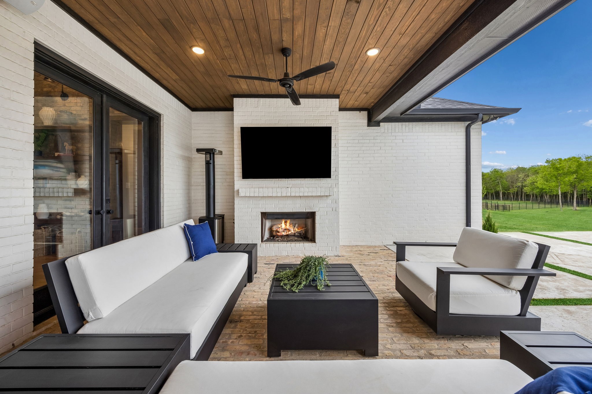 Covered patio with white-cushioned seating, a wood ceiling, and a lit fireplace overlooking the lawn.