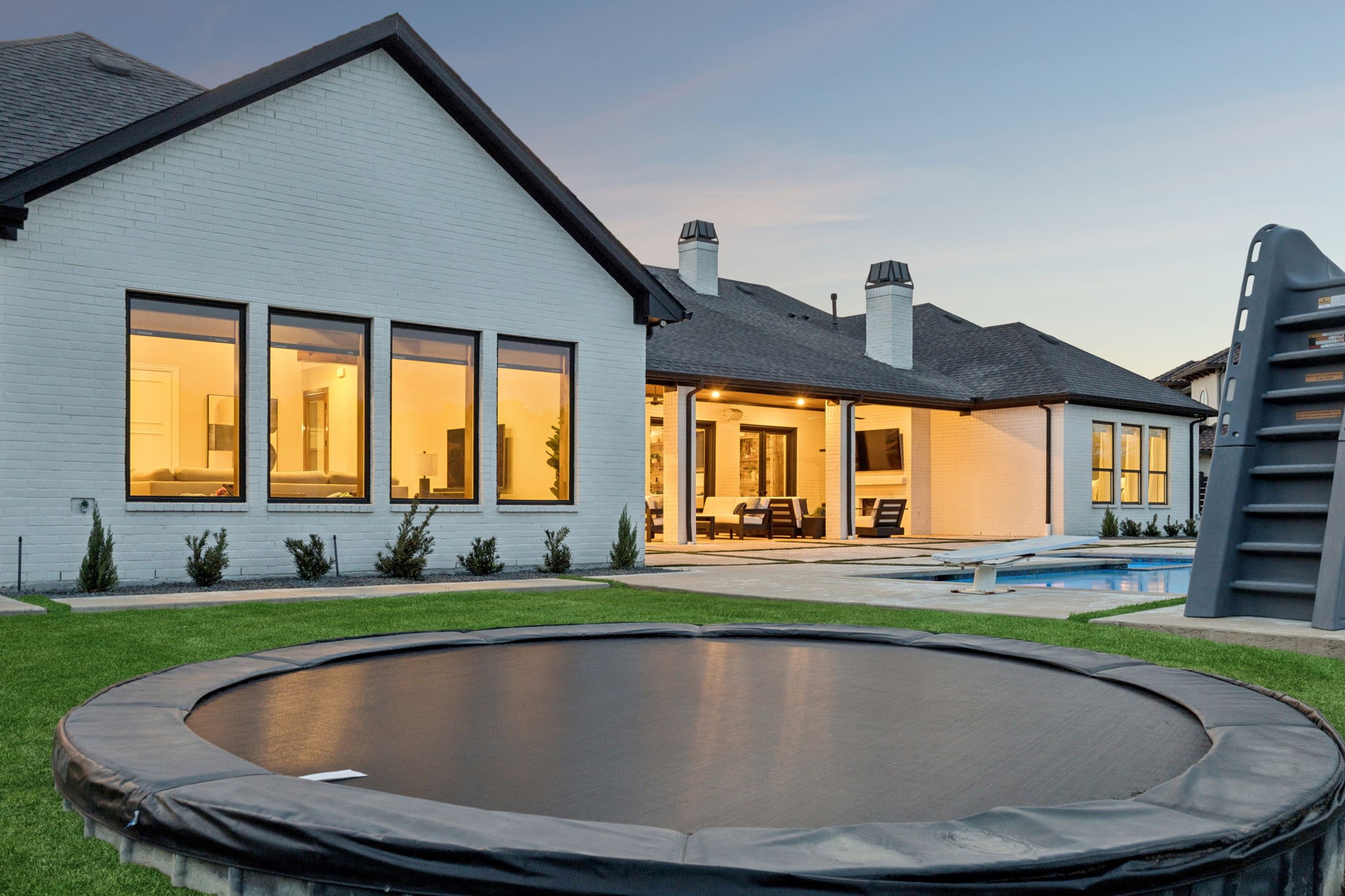 White-brick home with illuminated covered patio, swimming pool, and trampoline at dusk.