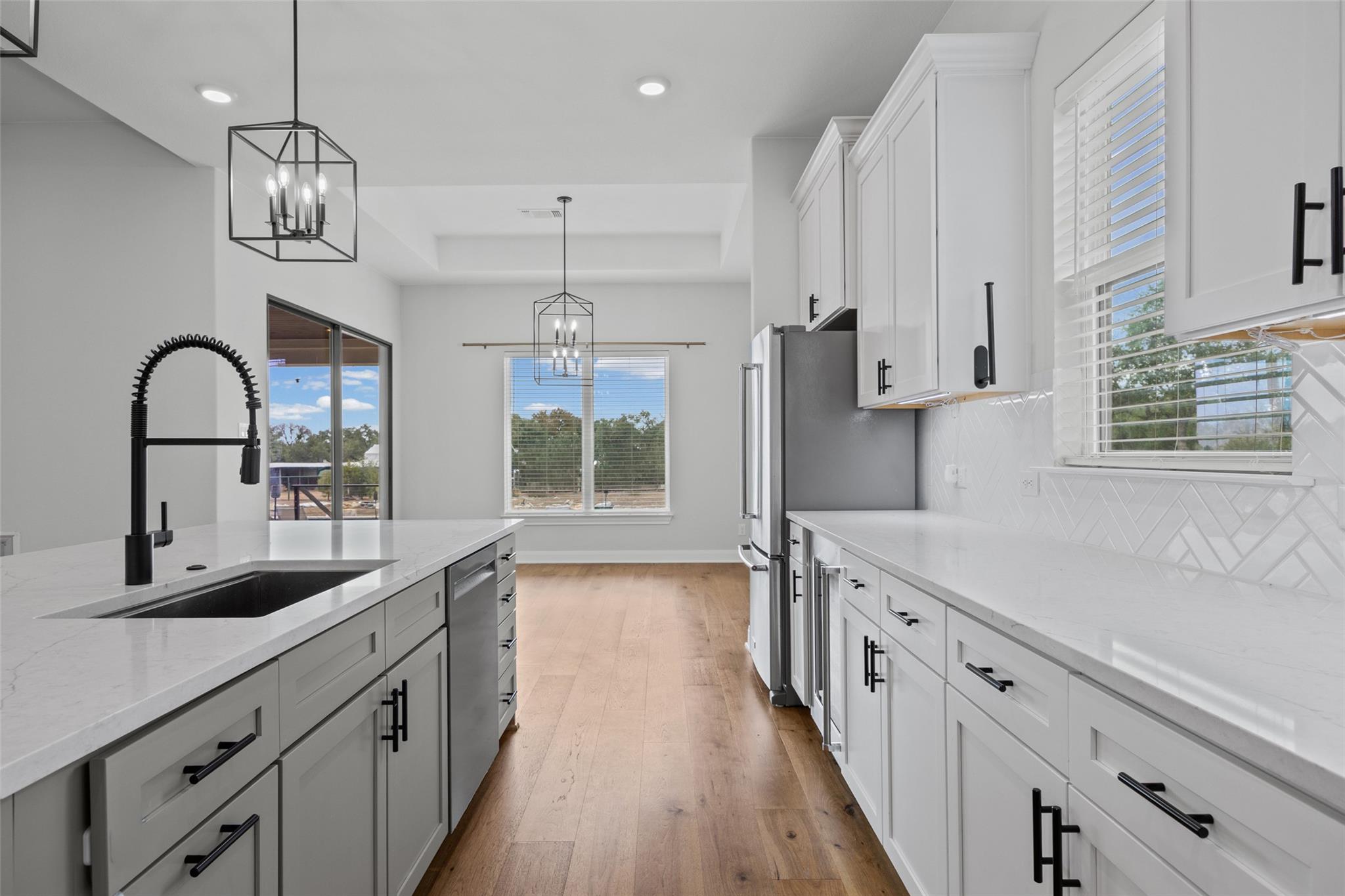 Modern white kitchen with a large island, black spring faucet, stainless refrigerator, and warm wood floors.