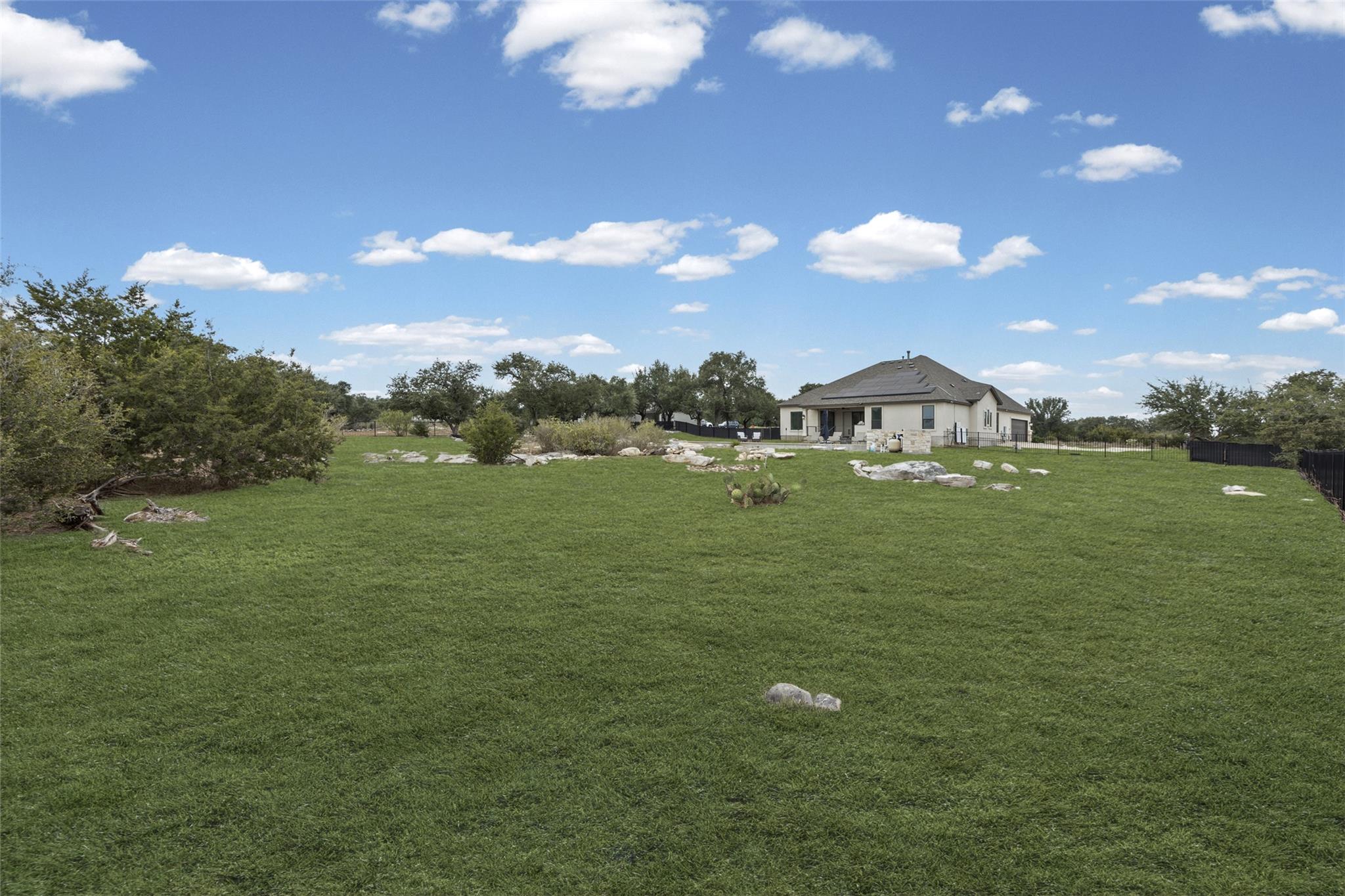 House beyond a broad grassy backyard with scattered rocks, mature trees, and black fencing.