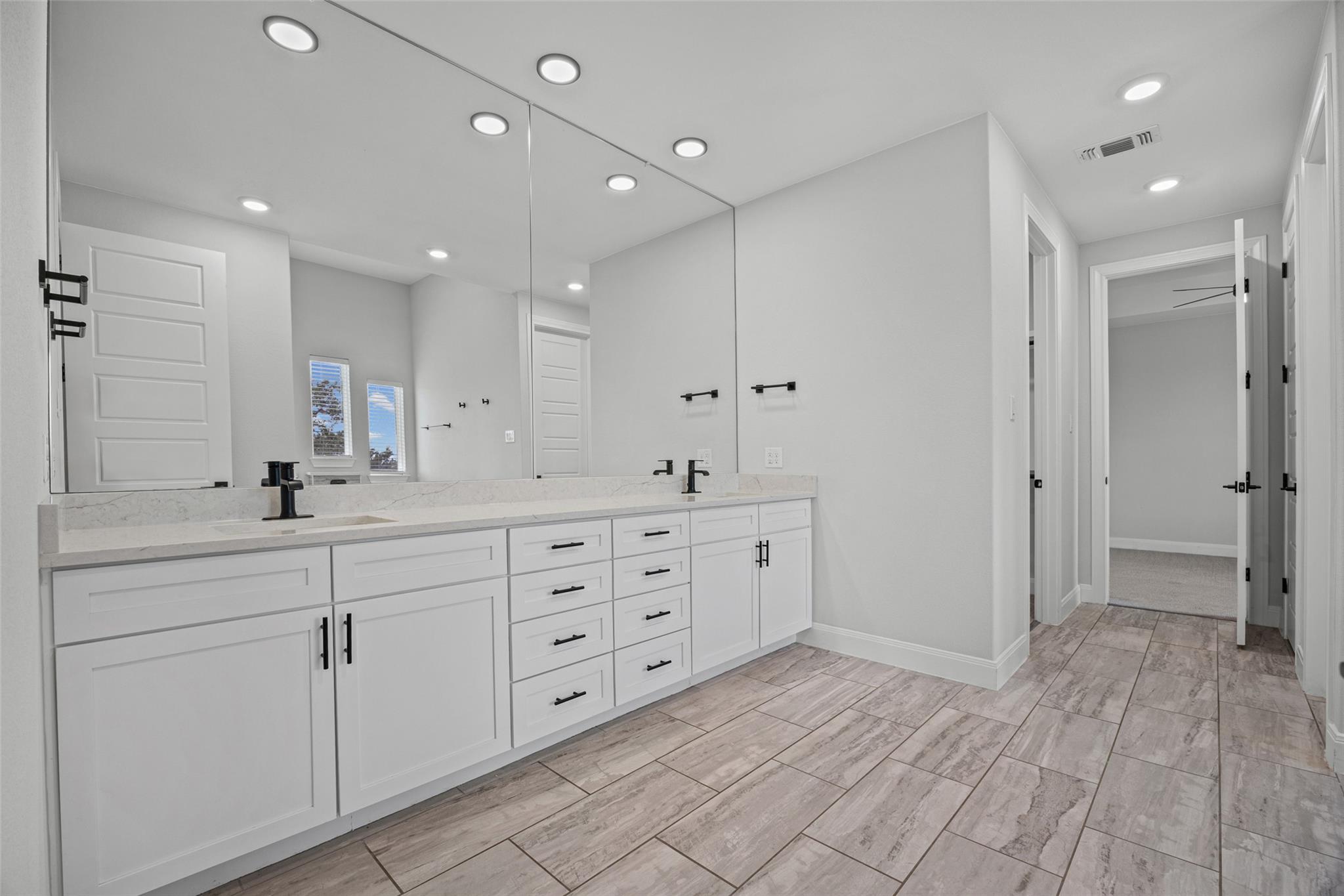Bathroom with a long white vanity, large mirror, black fixtures, and wood-look tile floor.