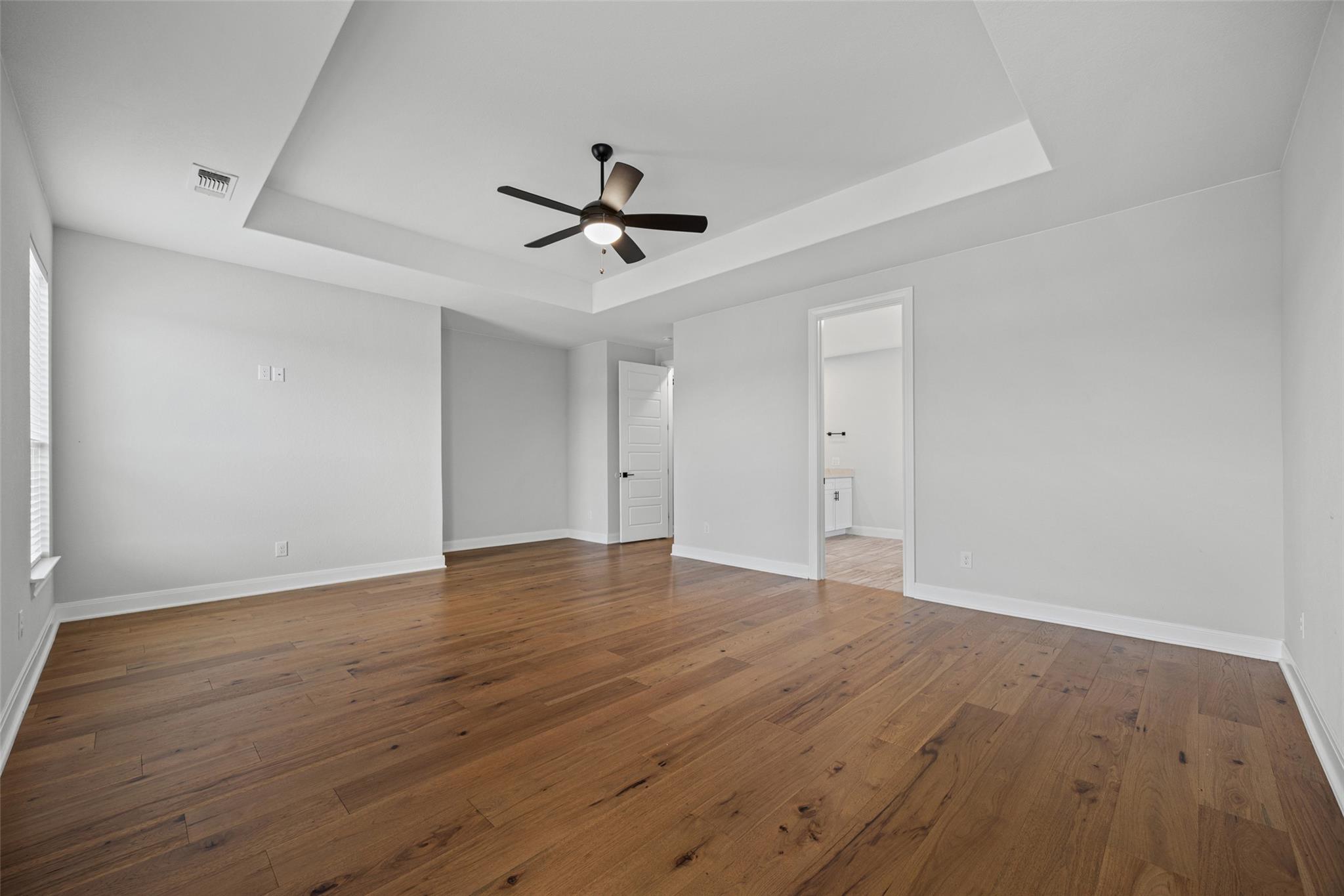 Empty bedroom with hardwood floors, a tray ceiling, ceiling fan, and doorway to a bathroom.