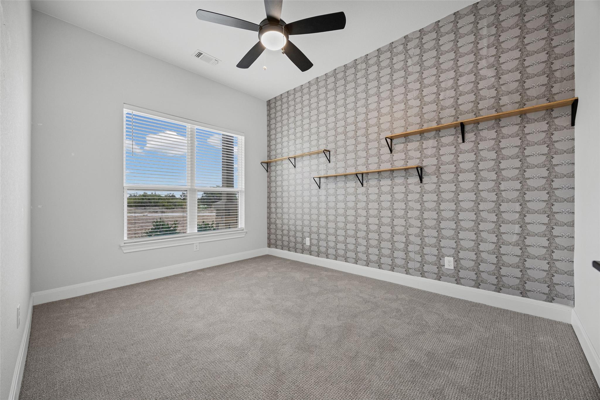 Empty bedroom with patterned wallpaper, floating shelves, a window, and a ceiling fan.